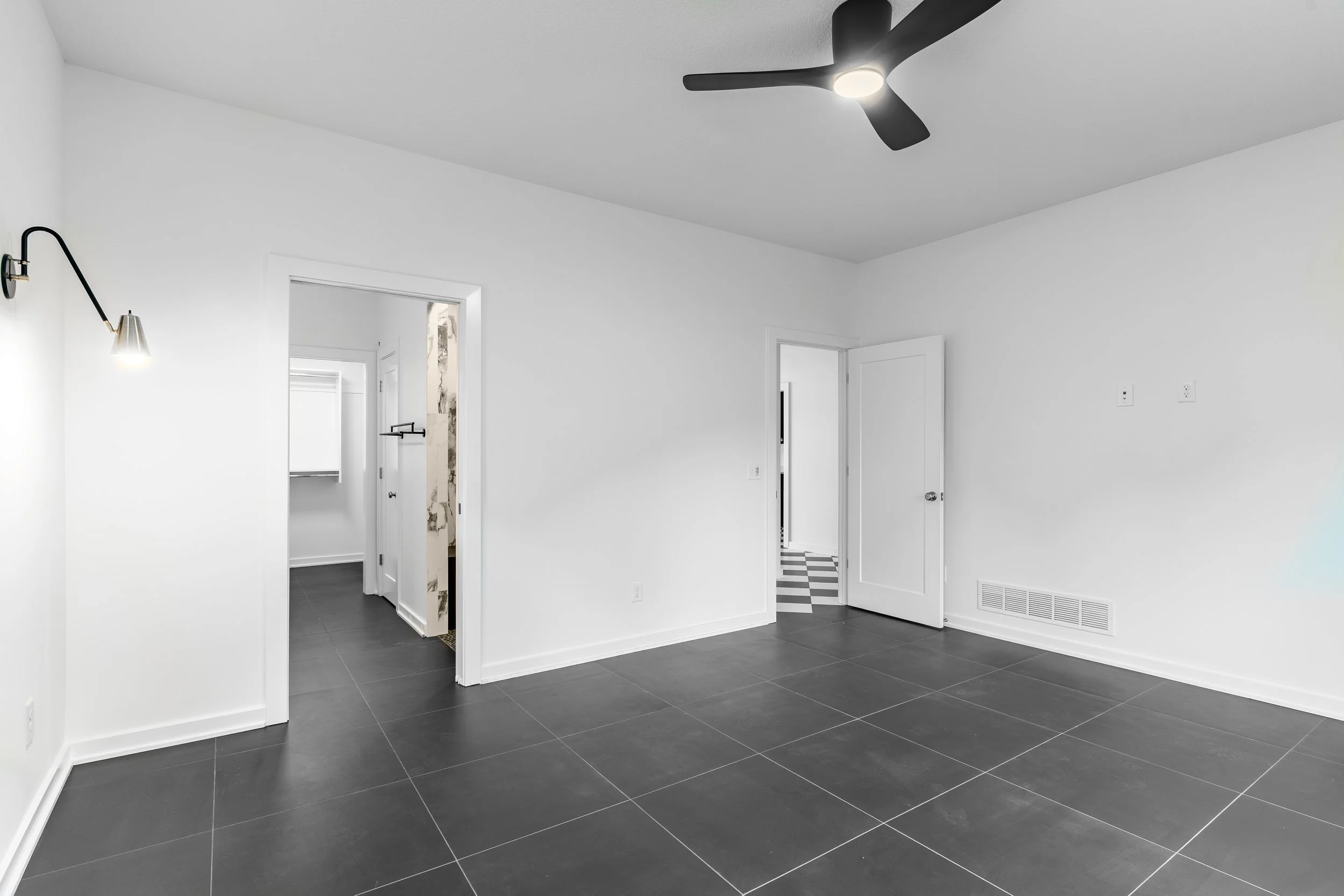 Empty room with white walls, black tiled floor, ceiling fan, wall-mounted light, and open doors leading to other rooms.