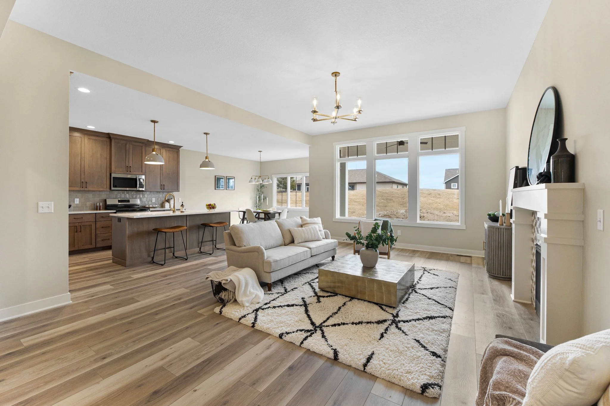 Open-concept living room and kitchen with large windows, beige walls, hardwood flooring, a white sofa, modern coffee table, area rug, and minimalistic decor.