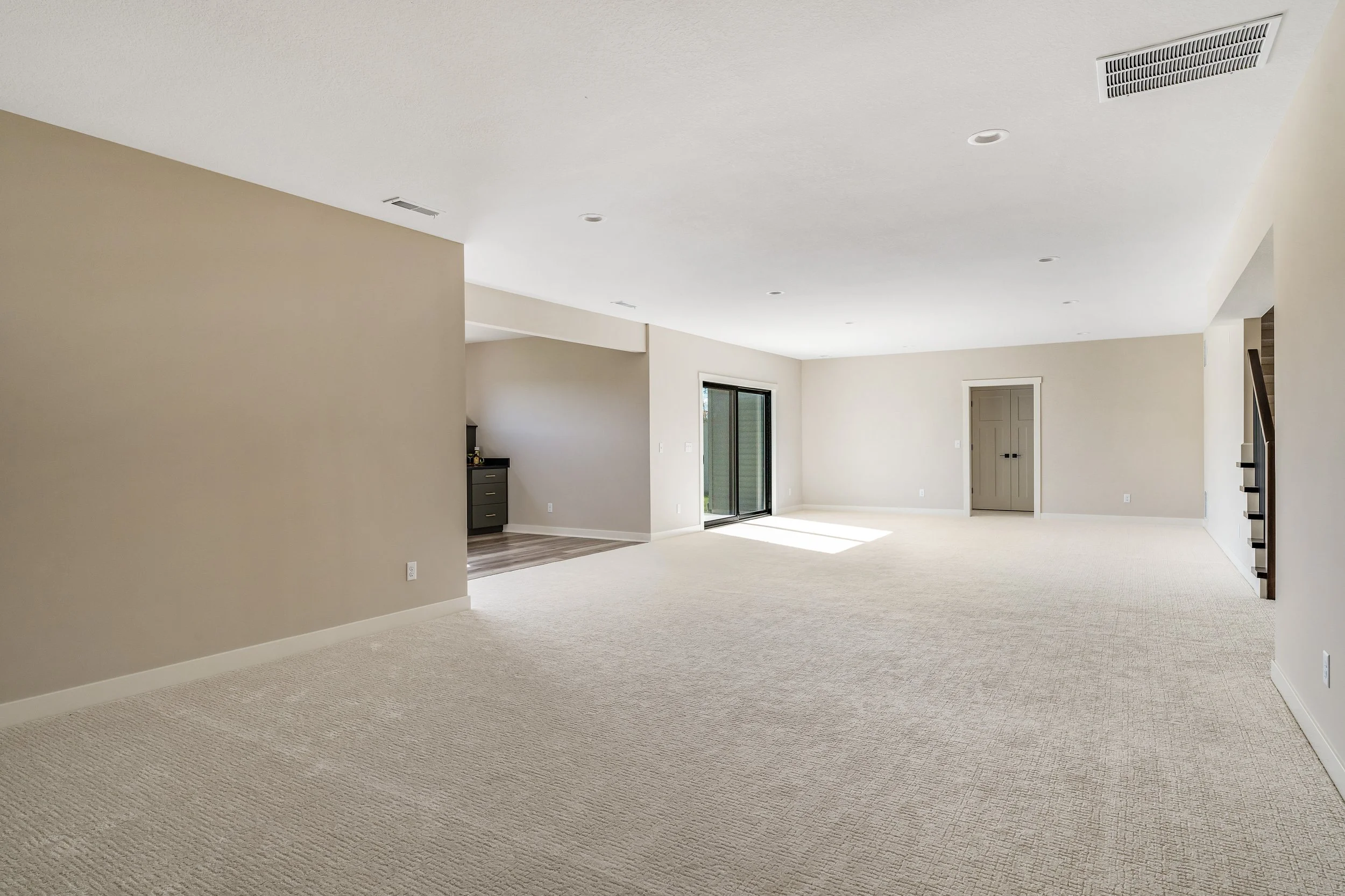 Empty living room with beige walls, white ceiling, carpeted floor, large sliding glass door, and staircase.