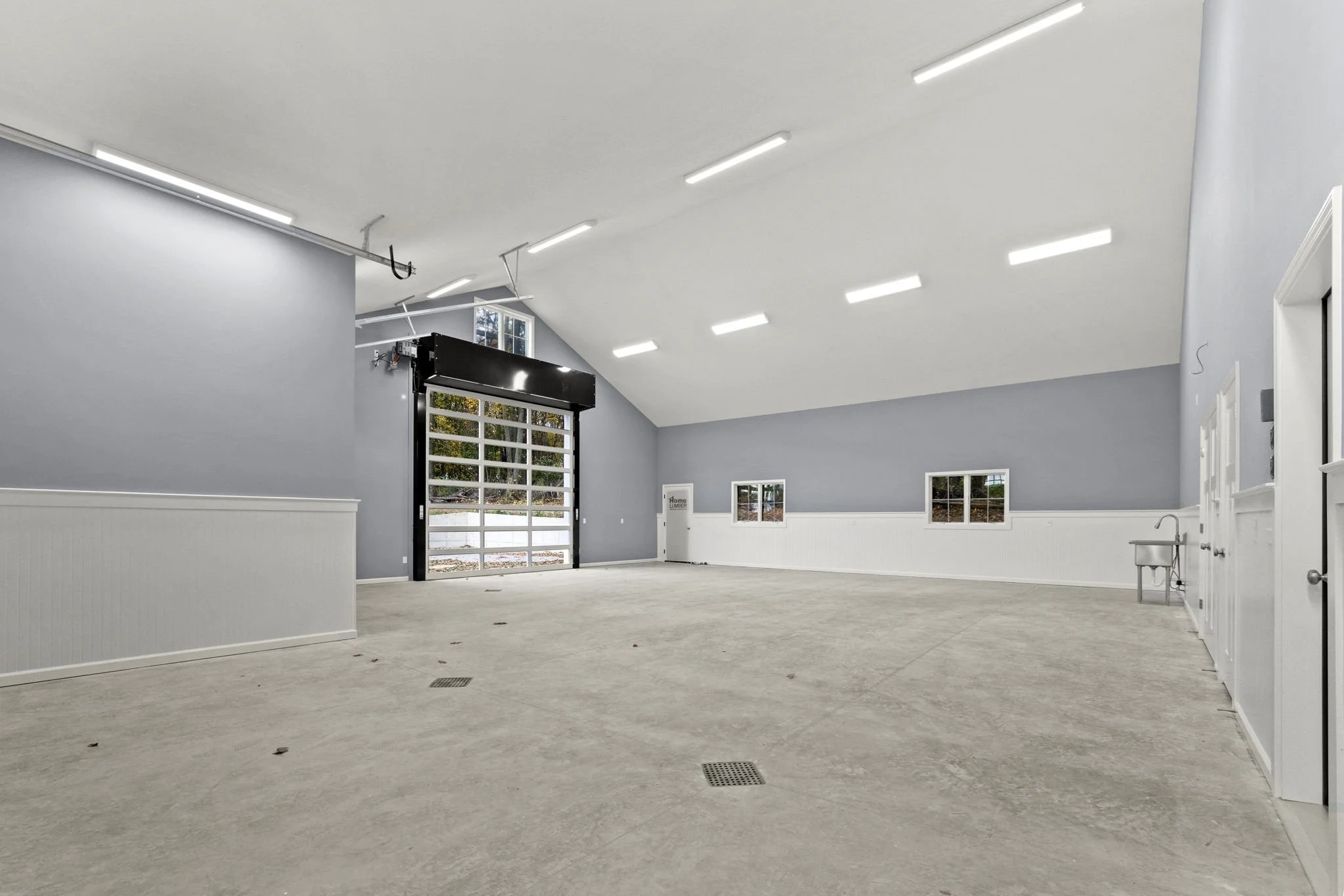 Empty garage with gray walls, white wainscoting, concrete floor, and bright ceiling lights, with a large garage door at the back and small windows on the right wall.
