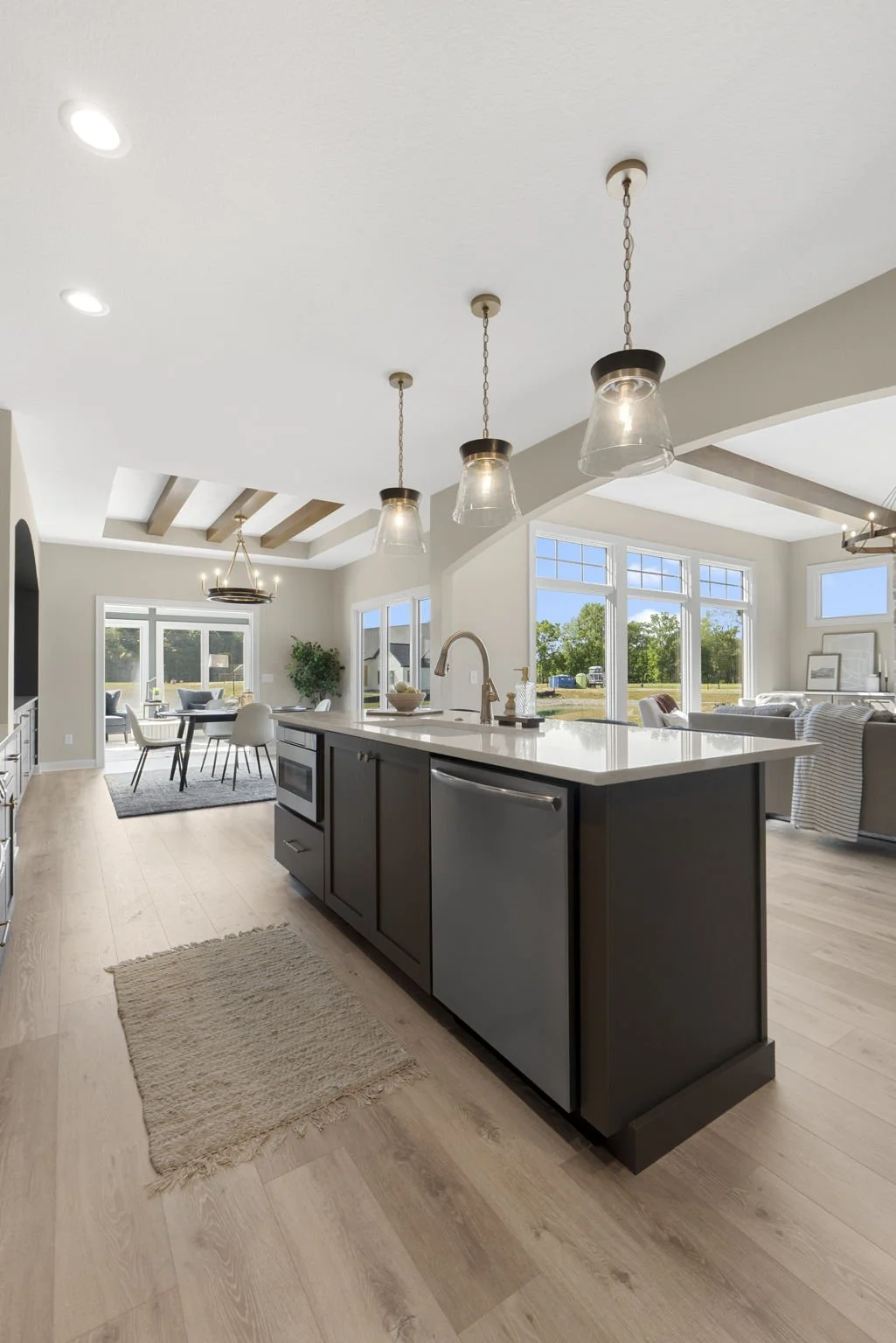 Open-concept kitchen and dining area with large windows, light wood flooring, a centered kitchen island with dark cabinets and a white countertop, pendant lights above, and a view of a living room and outside landscape.