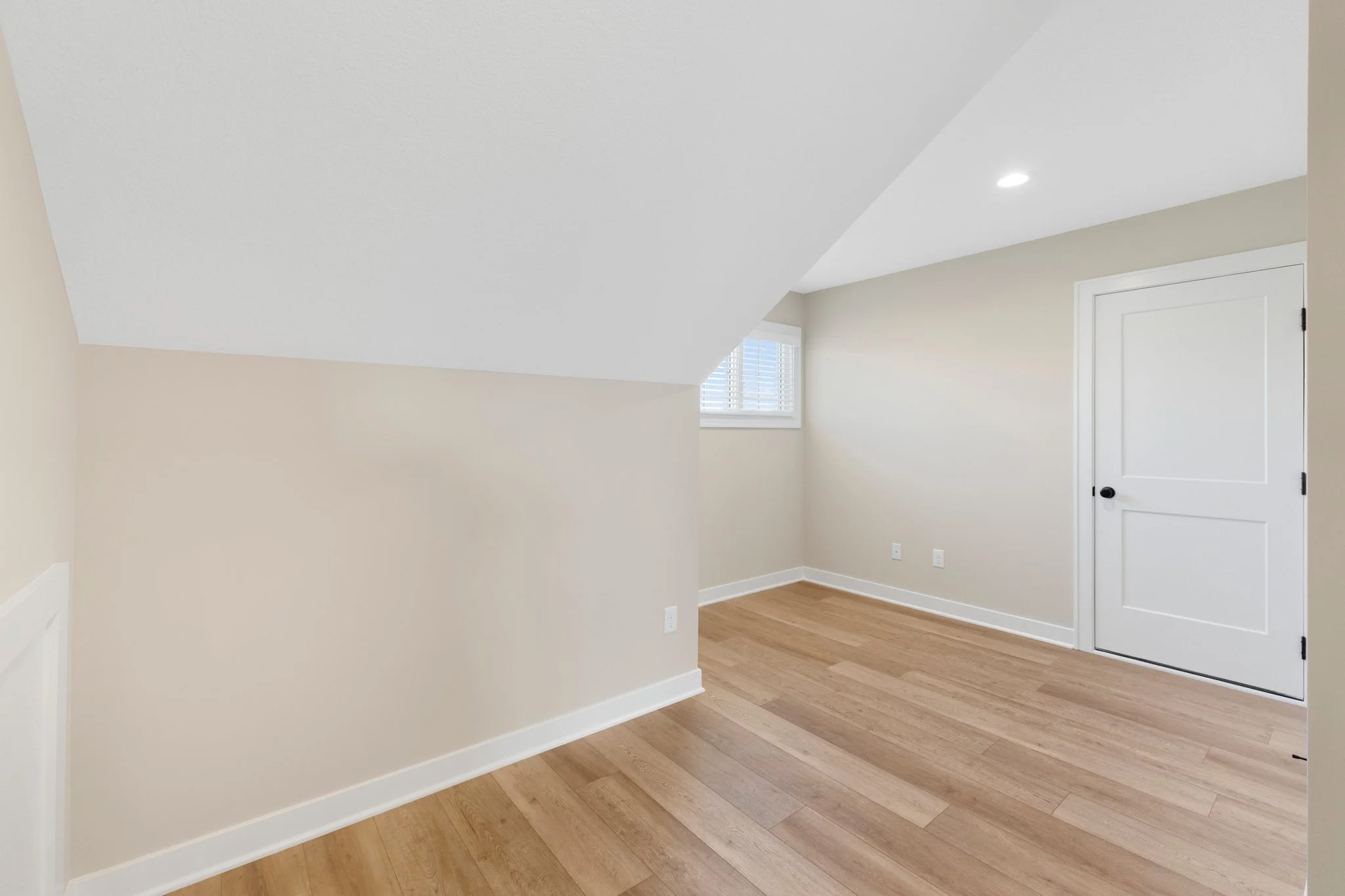 Empty room with angled ceiling, hardwood floors, small window with blinds, white door, and beige painted walls.