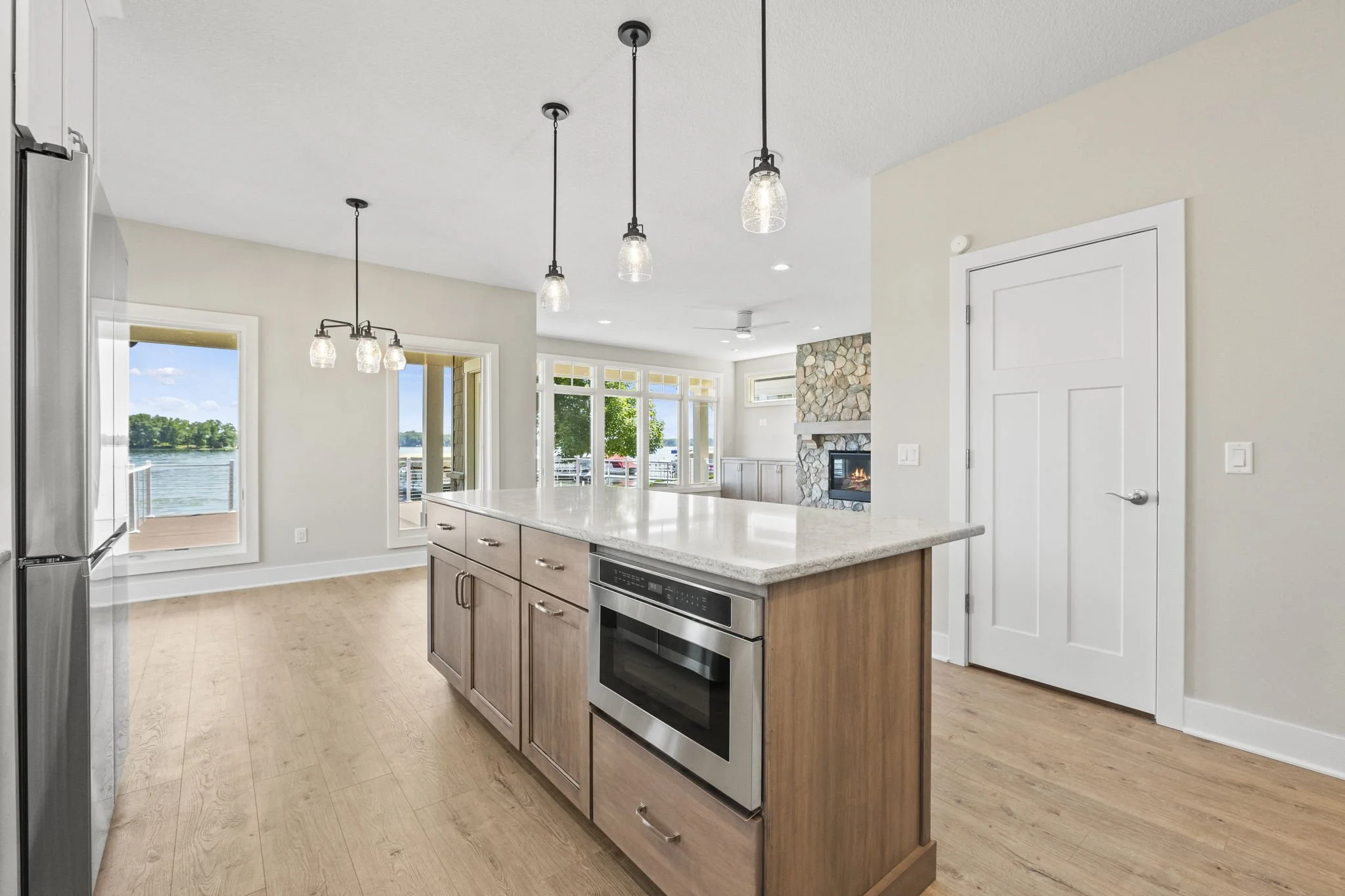 Modern kitchen with a large island with a light-colored granite countertop, wood cabinets, stainless steel oven, and hanging pendant lights, with large windows and a view of the water in the background.