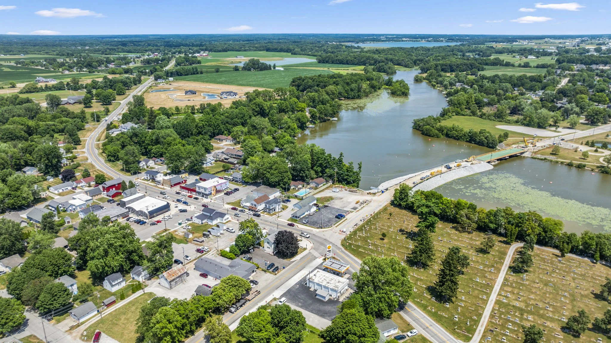 Aerial view of a small town with a river running through it, a bridge under construction, commercial buildings, residential houses, and green fields.