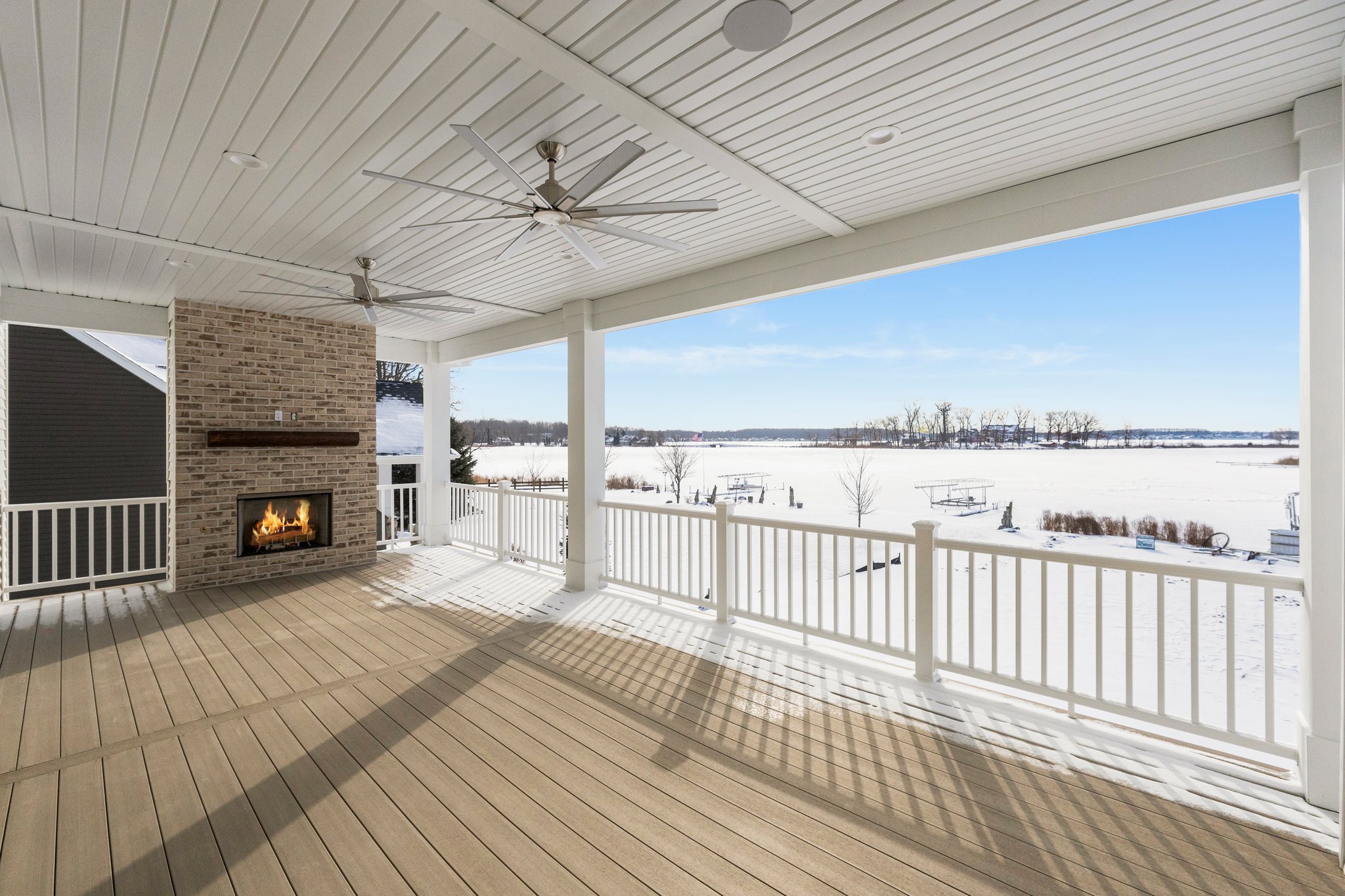 Covered porch overlooking snowy landscape with frozen lake and boats in the distance, brick fireplace and ceiling fans.