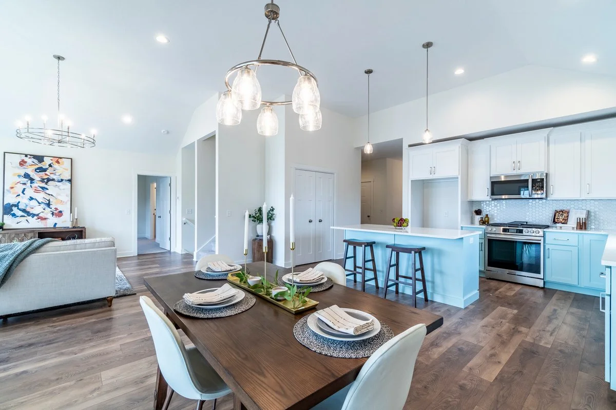 Open-concept living room and kitchen with white cabinetry, stainless steel appliances, a wooden dining table set for four, and modern lighting fixtures.