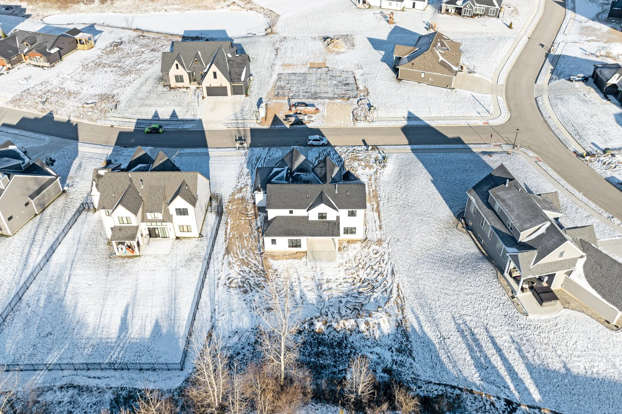 Aerial view of a snowy residential neighborhood with several houses, some under construction, and a street with cars casting long shadows.