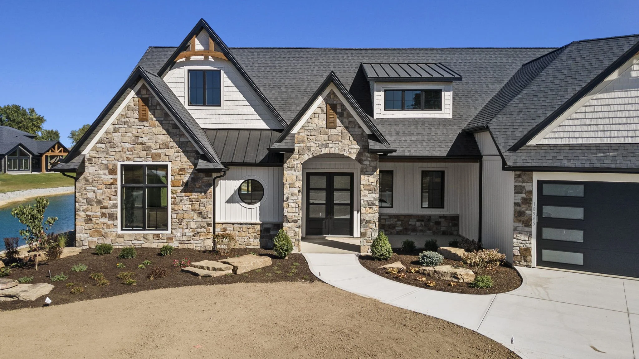 A modern house with a combination of stone, white siding, and black roof, featuring a curved walkway leading to the front door, with small landscaped plants and a bike lane in the background.