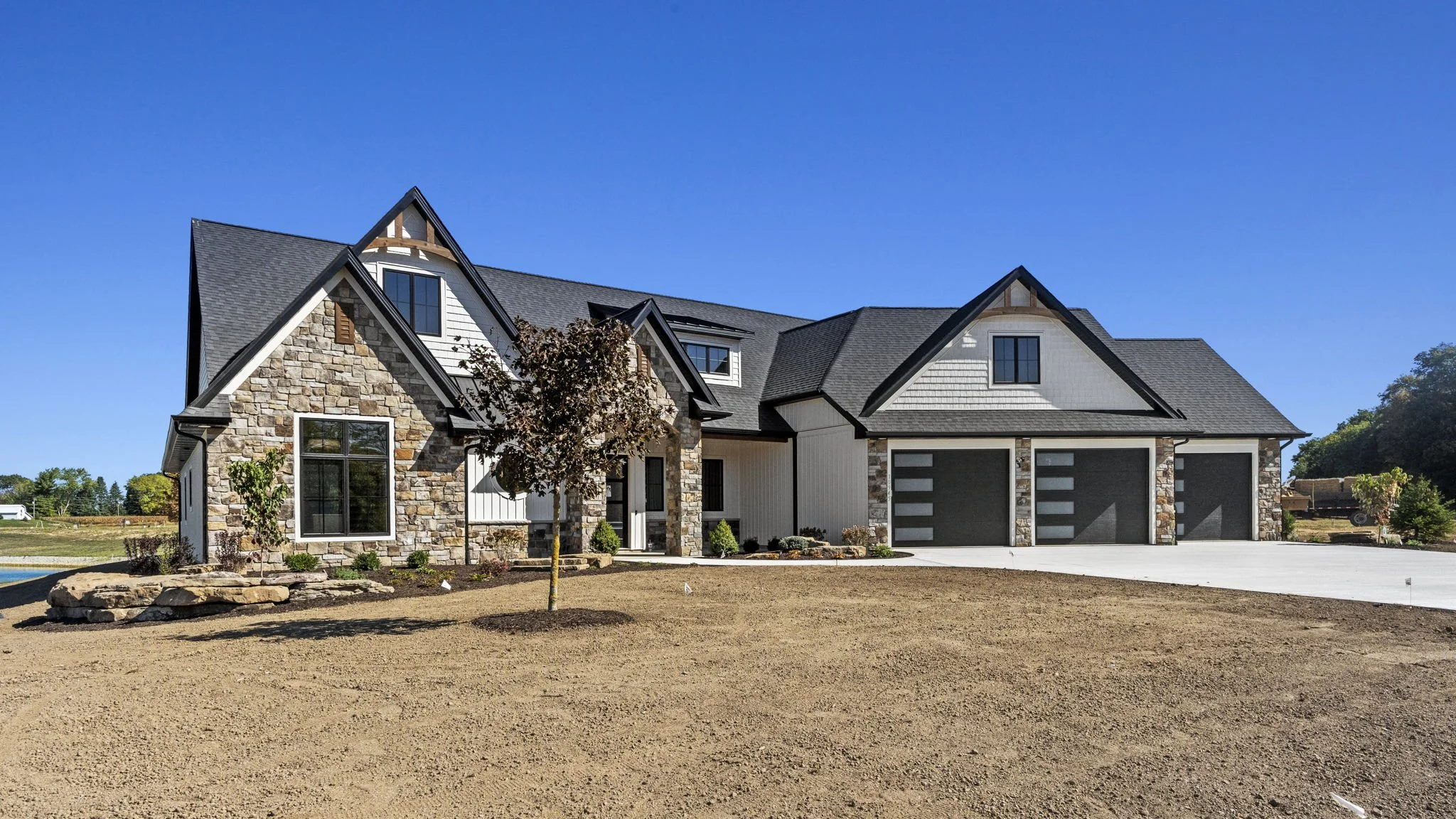 Newly constructed house with stone and white siding exterior, three garage doors, a small front yard with a tree, and a clear blue sky.