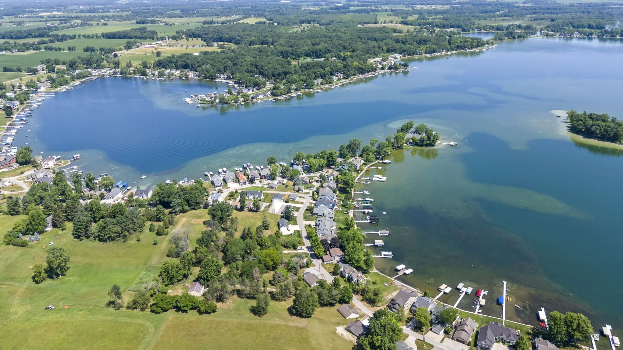 Aerial view of a lakeside neighborhood with houses, docks, boats, and green trees surrounding a large lake under a clear blue sky.