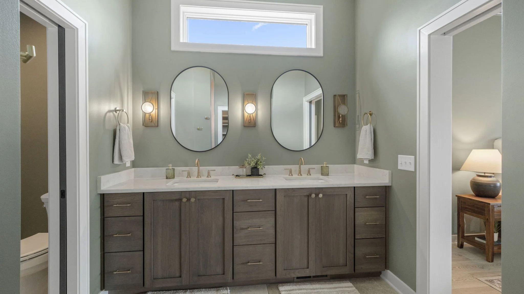 A modern bathroom with a double vanity, two oval mirrors, and a small window above. The vanity has a light wood finish with drawers and cabinet doors, and a white marble countertop with two sinks and gold fixtures. There are two wall-mounted light fi