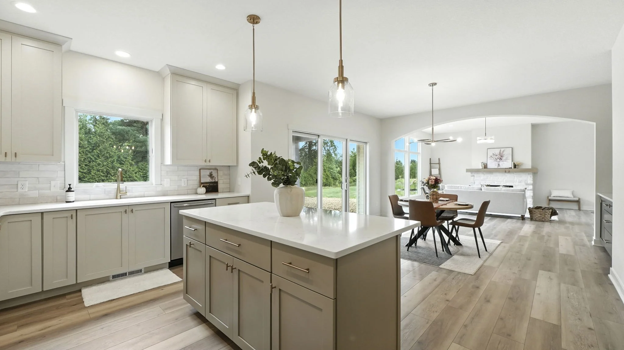 Open-concept kitchen and dining area with white cabinets, a kitchen island with a white countertop, hardwood floors, and large windows showing greenery outside.