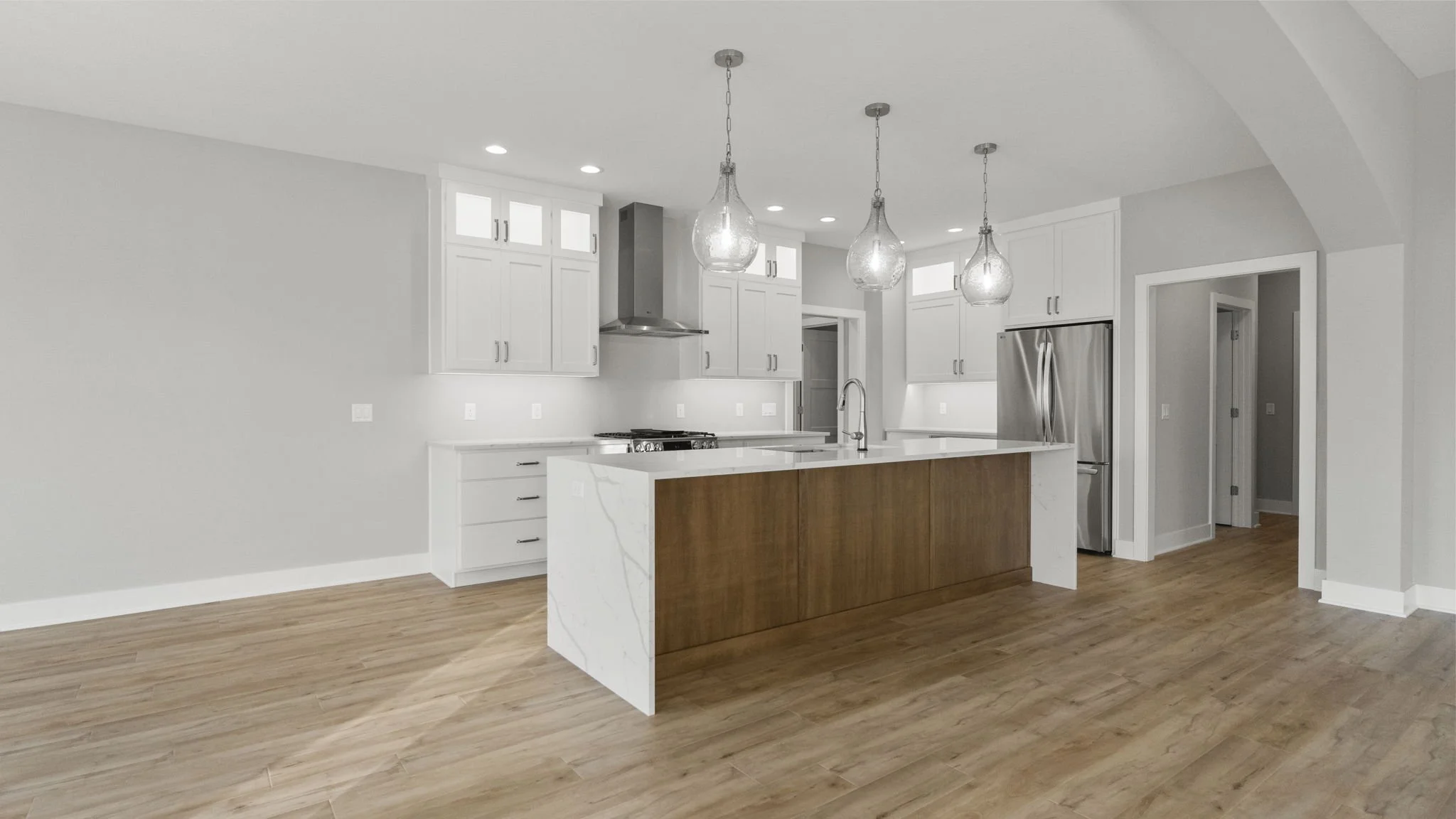 Modern kitchen with white cabinets, stainless steel refrigerator, island with wooden panels, light wood flooring, three pendant lights, and spotlights on the ceiling.