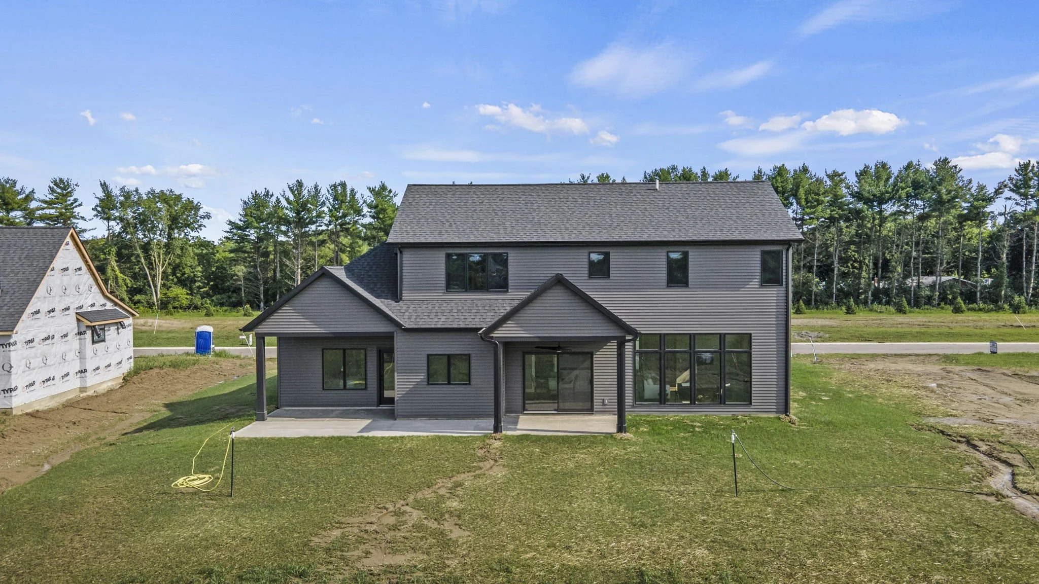 A new two-story gray house under construction with a porch and large windows, surrounded by a grassy yard and neighboring houses in the background, with a blue sky and some clouds.