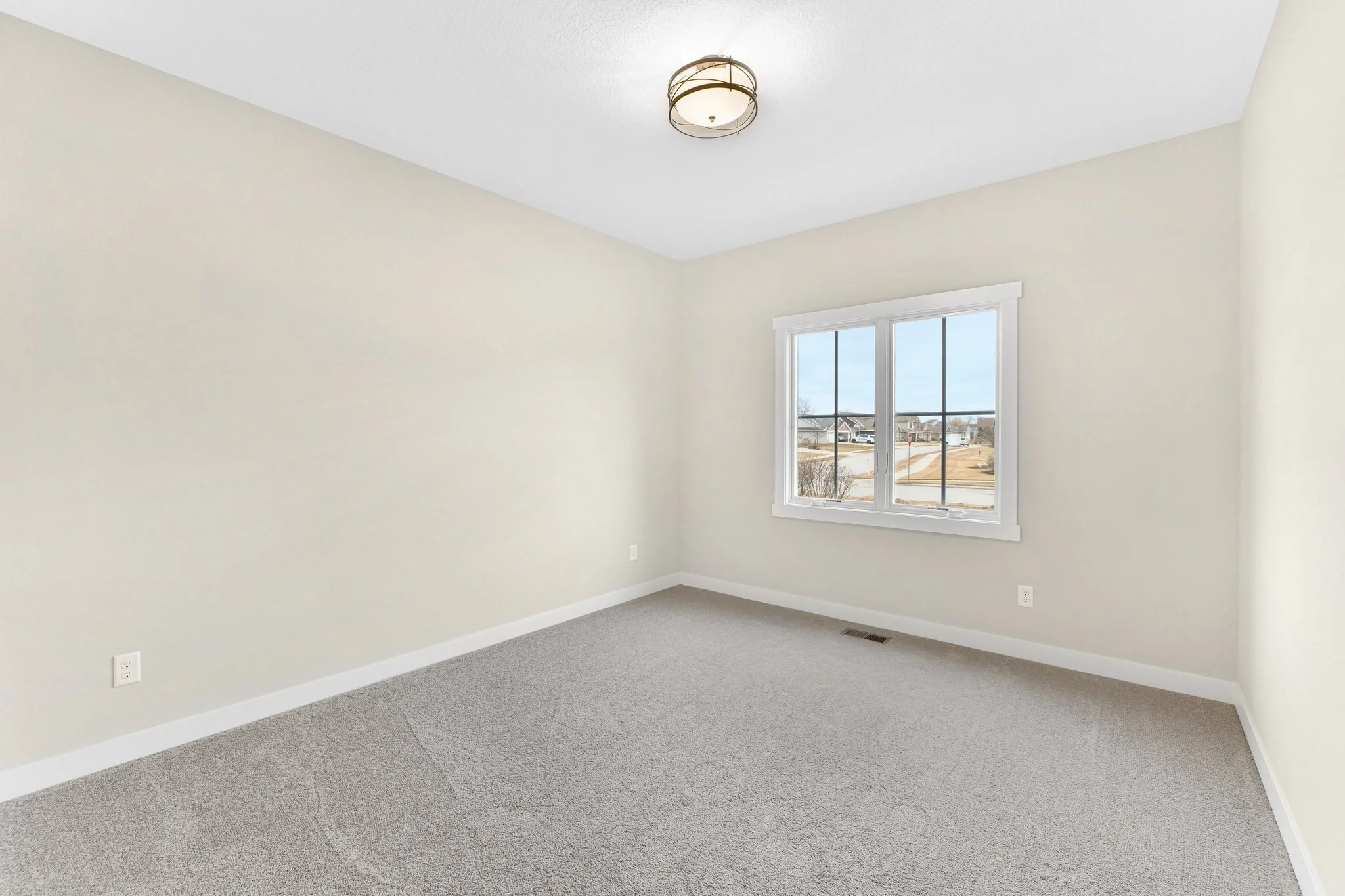Empty room with beige walls, gray carpet, white trim, a window showing a neighborhood, and a ceiling light fixture.