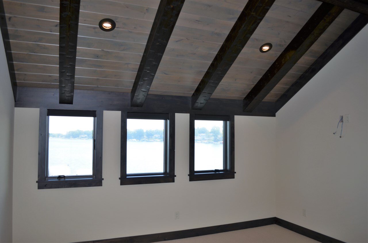Empty room with three windows, sloped ceiling with exposed dark wooden beams, beige walls, and black trim.
