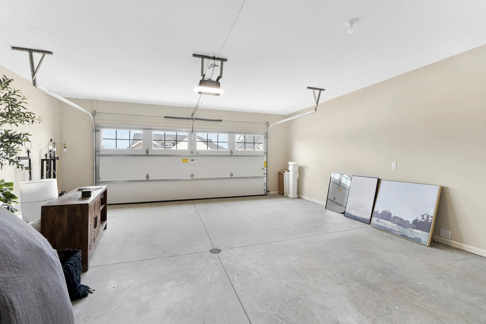 Empty garage with a white sectional garage door, a small wooden table, a tall green plant, and three framed pictures leaning against the beige wall.