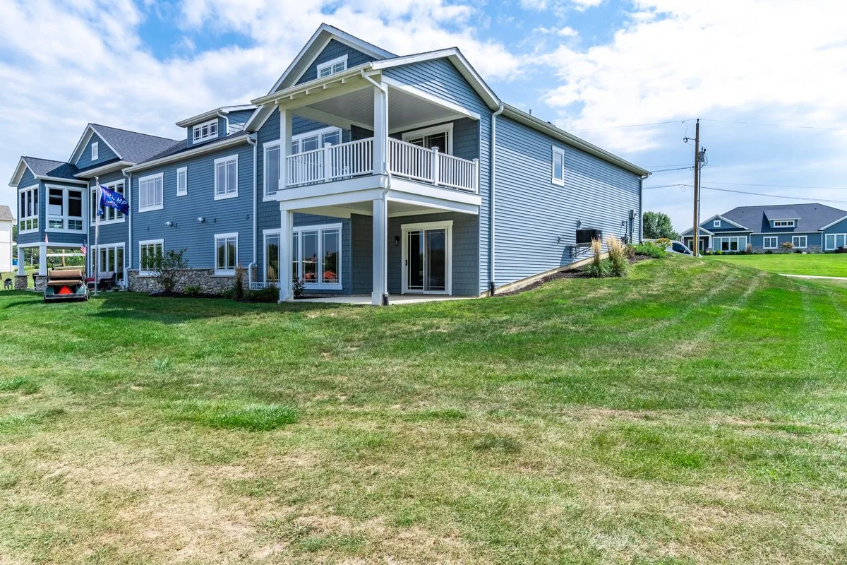 Blue multi-story house with white trim, balcony, and sliding glass doors, surrounded by green lawn and some neighboring houses in the background.