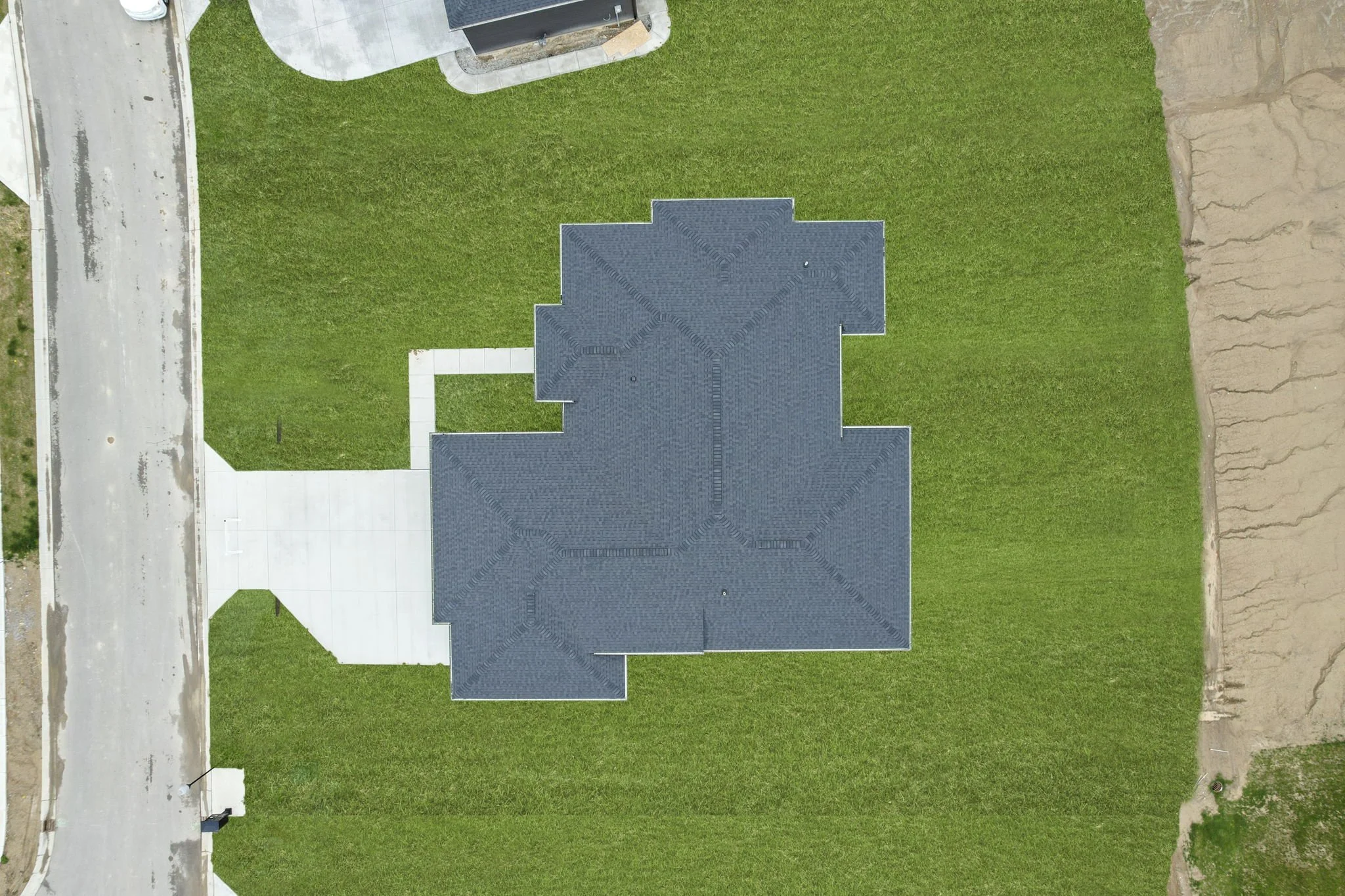 An aerial view of a house with a dark gray roof, surrounded by a green lawn, with a driveway leading to the front entrance and a sidewalk connecting the driveway to the street.