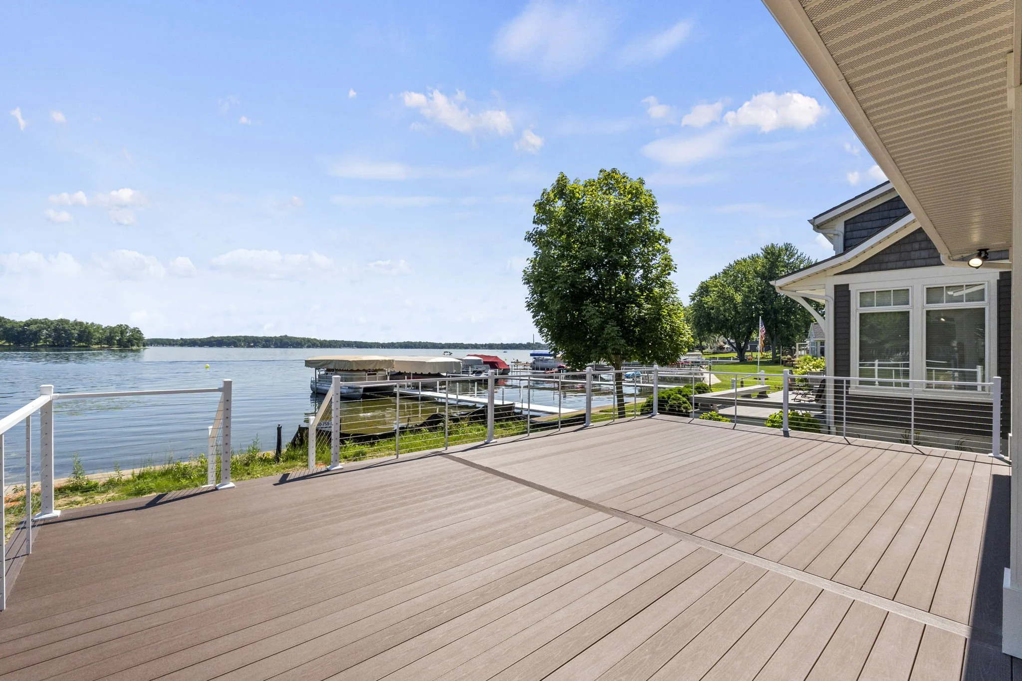 Large wooden deck overlooking a calm lake with boats moored by a dock, surrounded by a metal railing, under a partly cloudy sky.