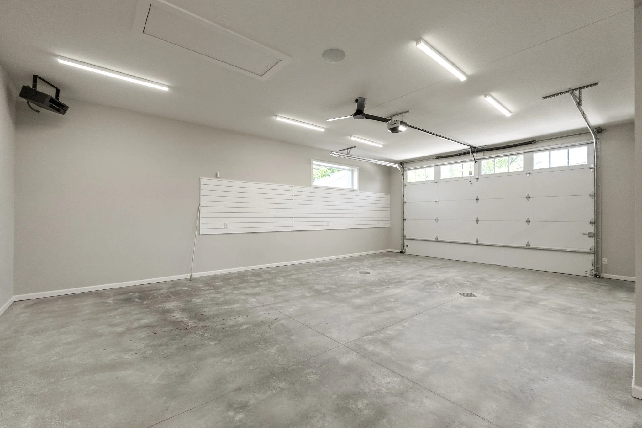 Empty clean garage with concrete floor, white walls, a large white garage door, rectangular overhead lights, and a small window, with a ceiling fan and a mounted garage door opener.