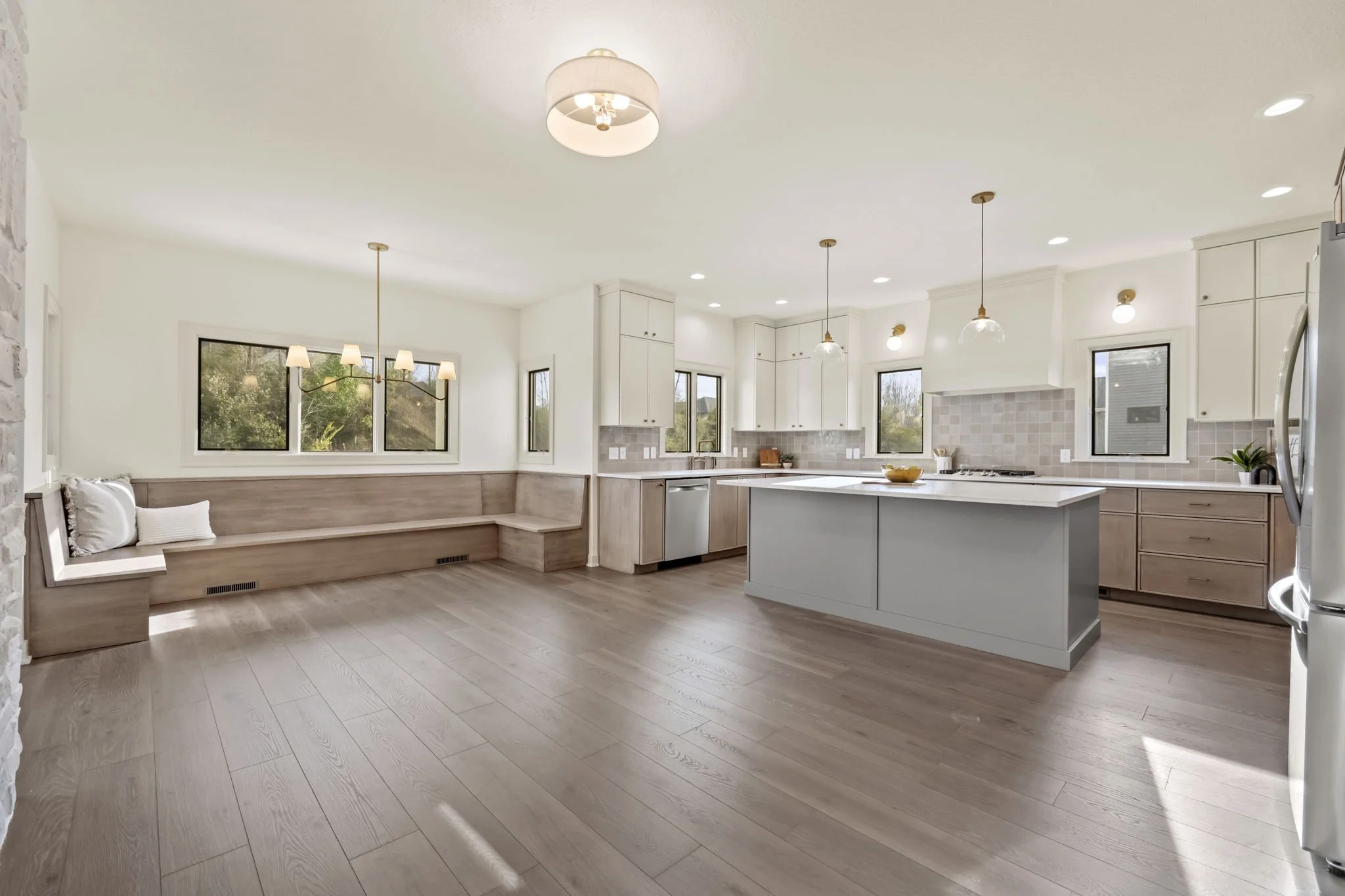 Open kitchen and dining area with light wood floors, white and light wood cabinetry, pendant lights over island, and a built-in bench with cushions along the wall, large windows letting in natural light.