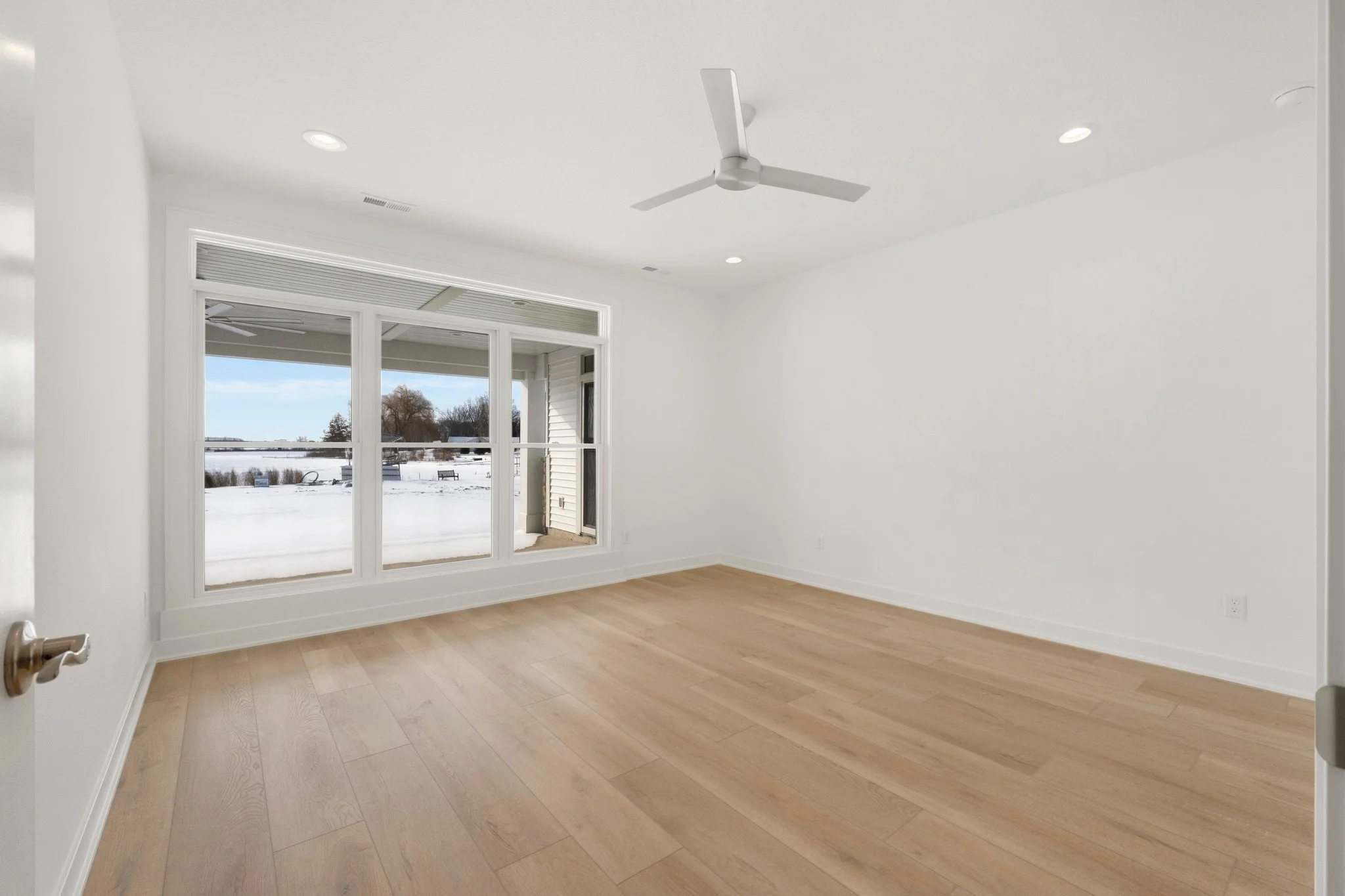 Empty room with white walls, large window showing snowy outdoor scene, ceiling fan, and light wood flooring.