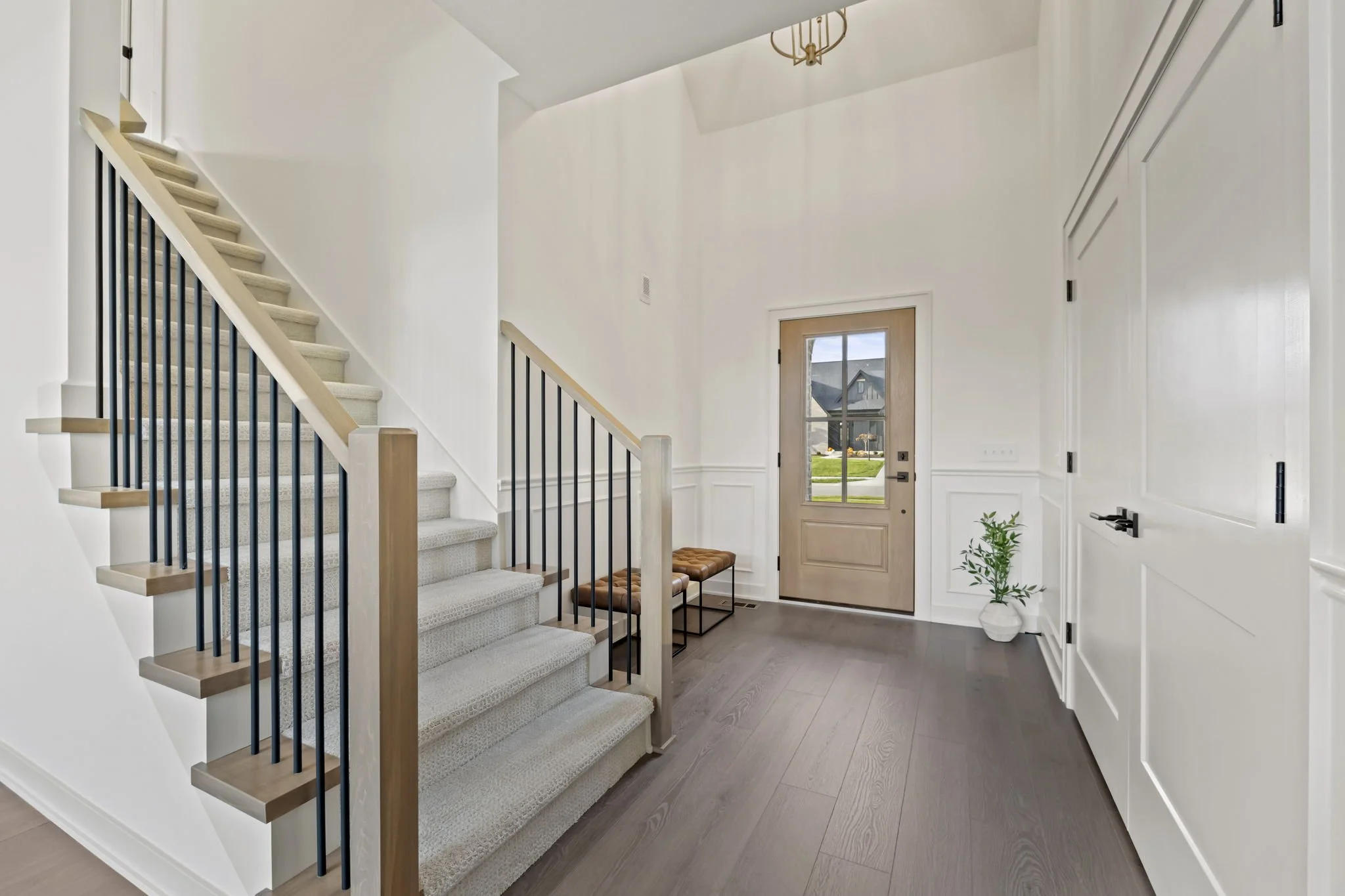 Modern entryway with a staircase featuring wooden steps and black metal railing, a wooden bench with leather cushions, a wood front door with glass window, a small potted plant, and dark hardwood flooring.