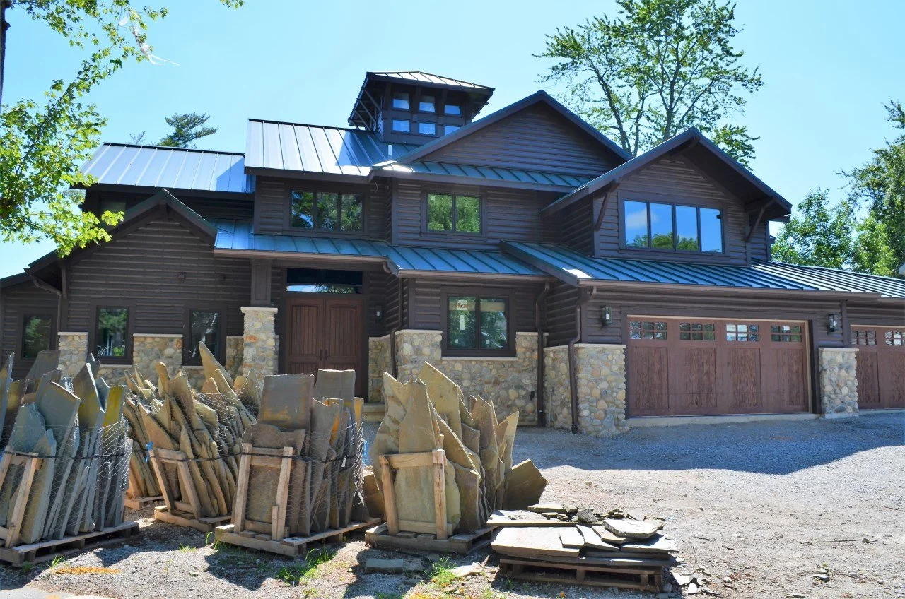 A large modern house with dark wood siding, stone accents, and a metal roof, surrounded by blue sky and trees.