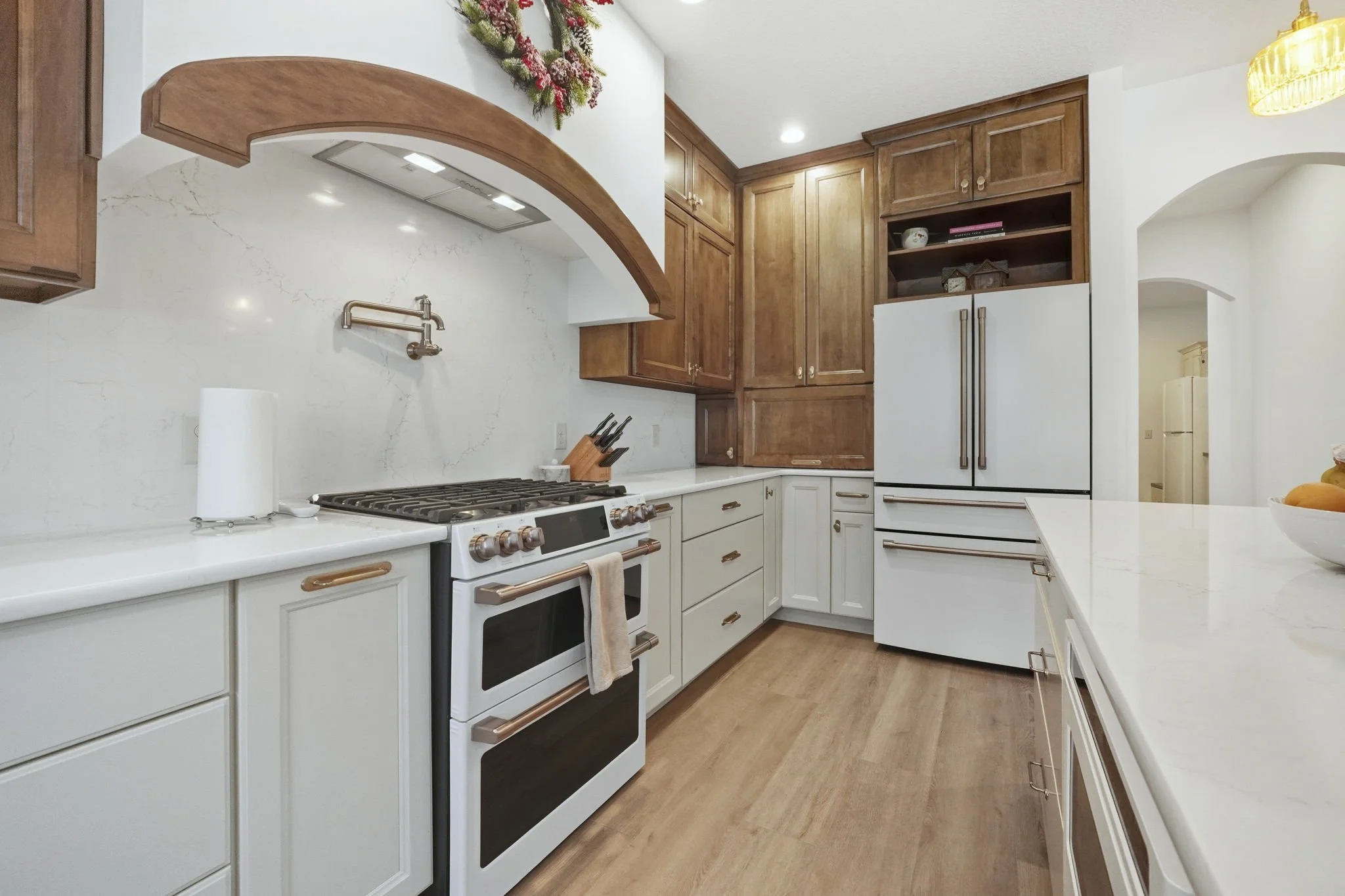 A kitchen with white cabinets, a white stove with copper handles, and a wooden ceiling arch. There is a paper towel roll on the counter, a knife block near the stove, and a large white refrigerator. The kitchen has light wood floors and a decorative 