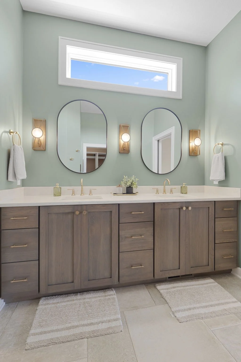 A double sink bathroom vanity with light wood cabinets, two oval mirrors, and gold fixtures, with a window above the mirrors and two wall-mounted light fixtures.