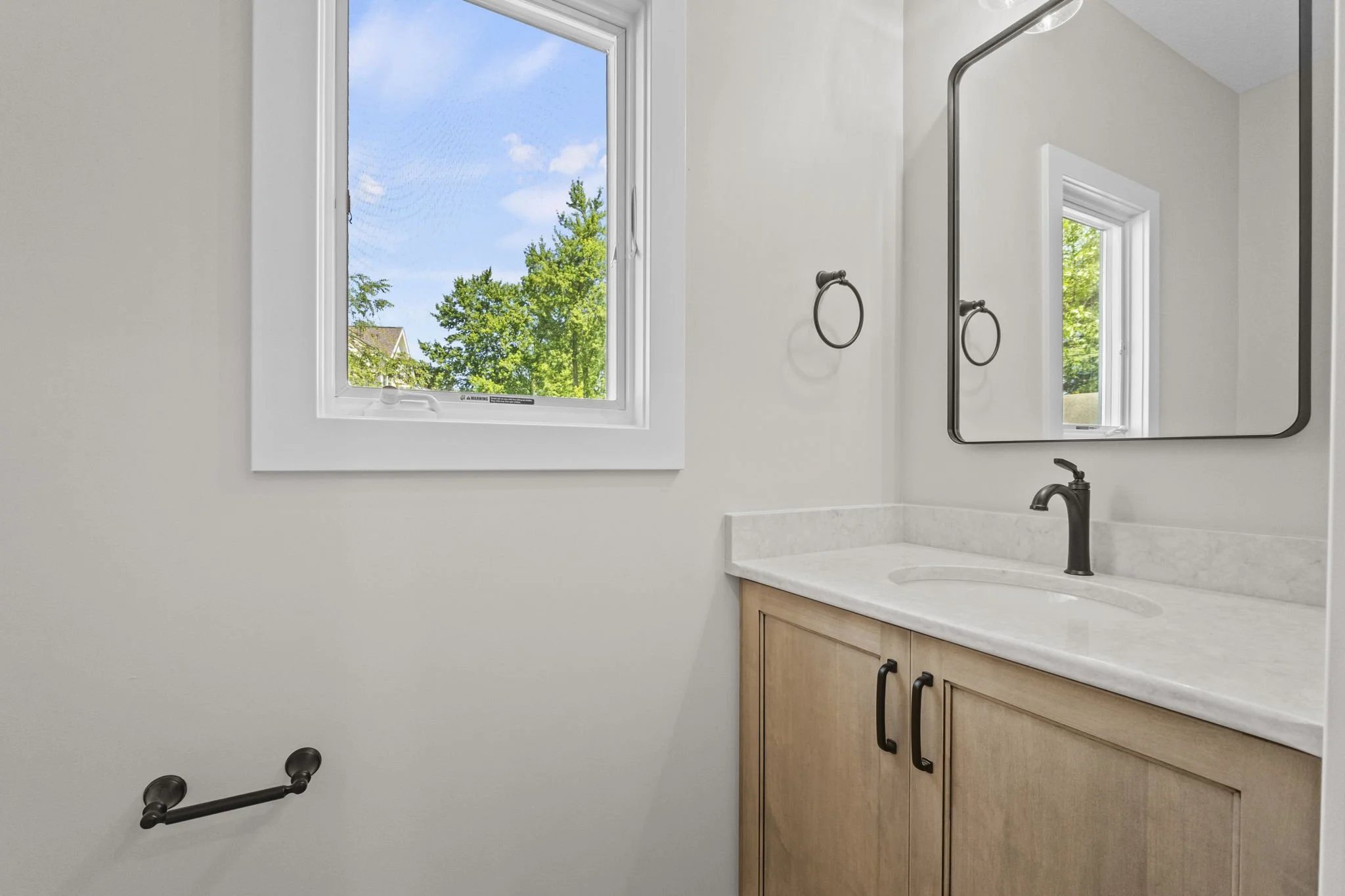 A bathroom vanity with a light wooden cabinet, a white marble countertop, and a black faucet. There is a large mirror above the sink, with two black towel rings on the wall. A window shows trees and a blue sky outside.