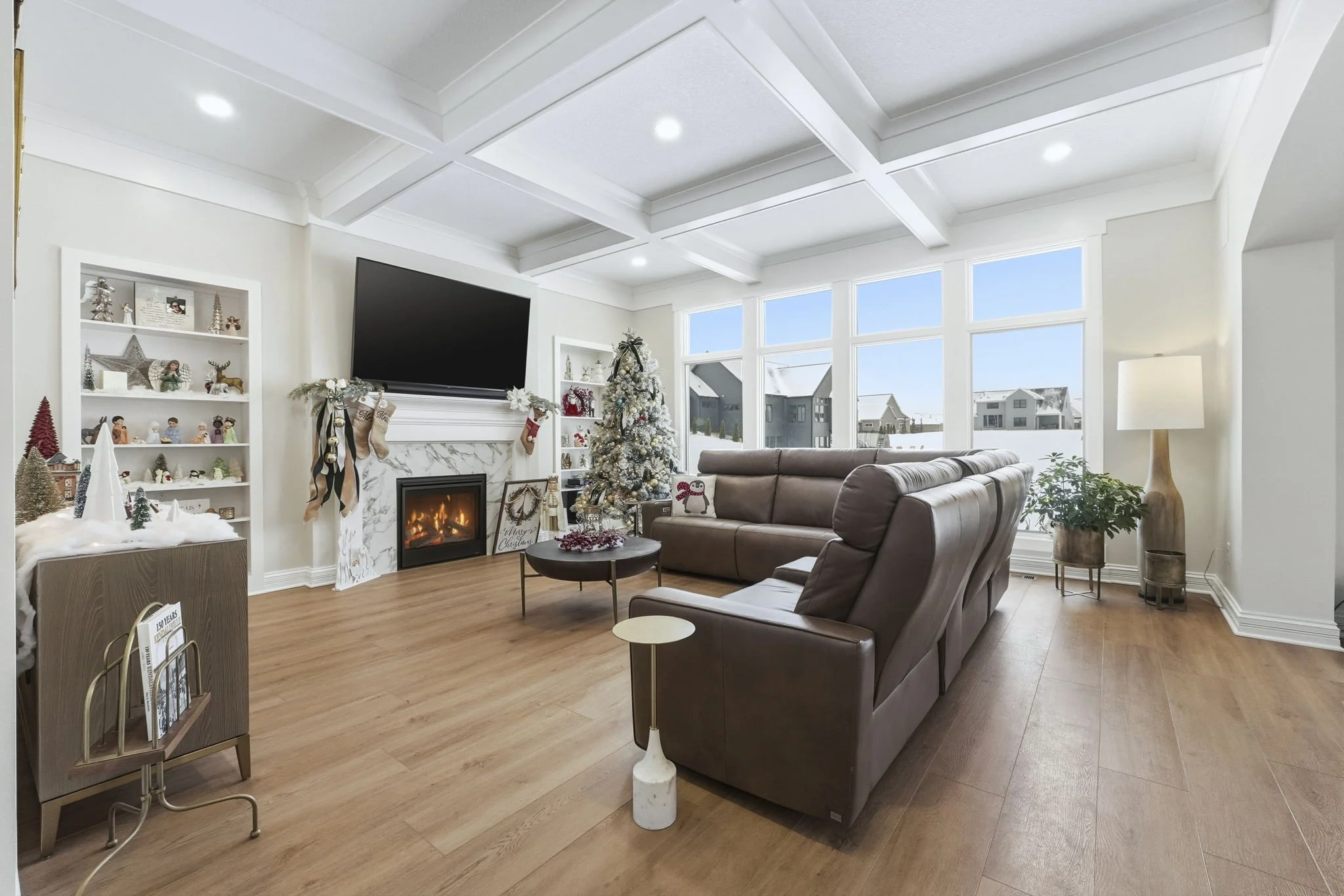 Living room decorated for Christmas with a white Christmas tree, stockings on the fireplace, and holiday decorations on the shelves and tables, with large windows showing a snowy neighborhood outside.