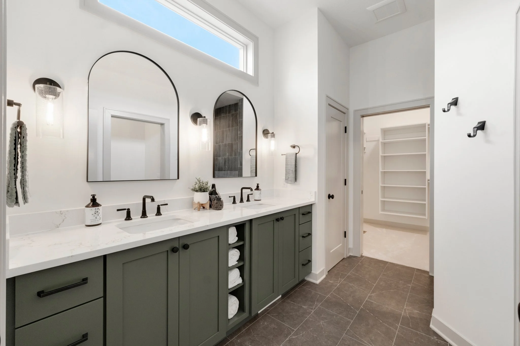 Modern bathroom with double vanity, green cabinets, white marble countertop, two large arched mirrors, black faucet fixtures, large window for natural light, gray tiled floor, and a walk-in closet visible in the background.