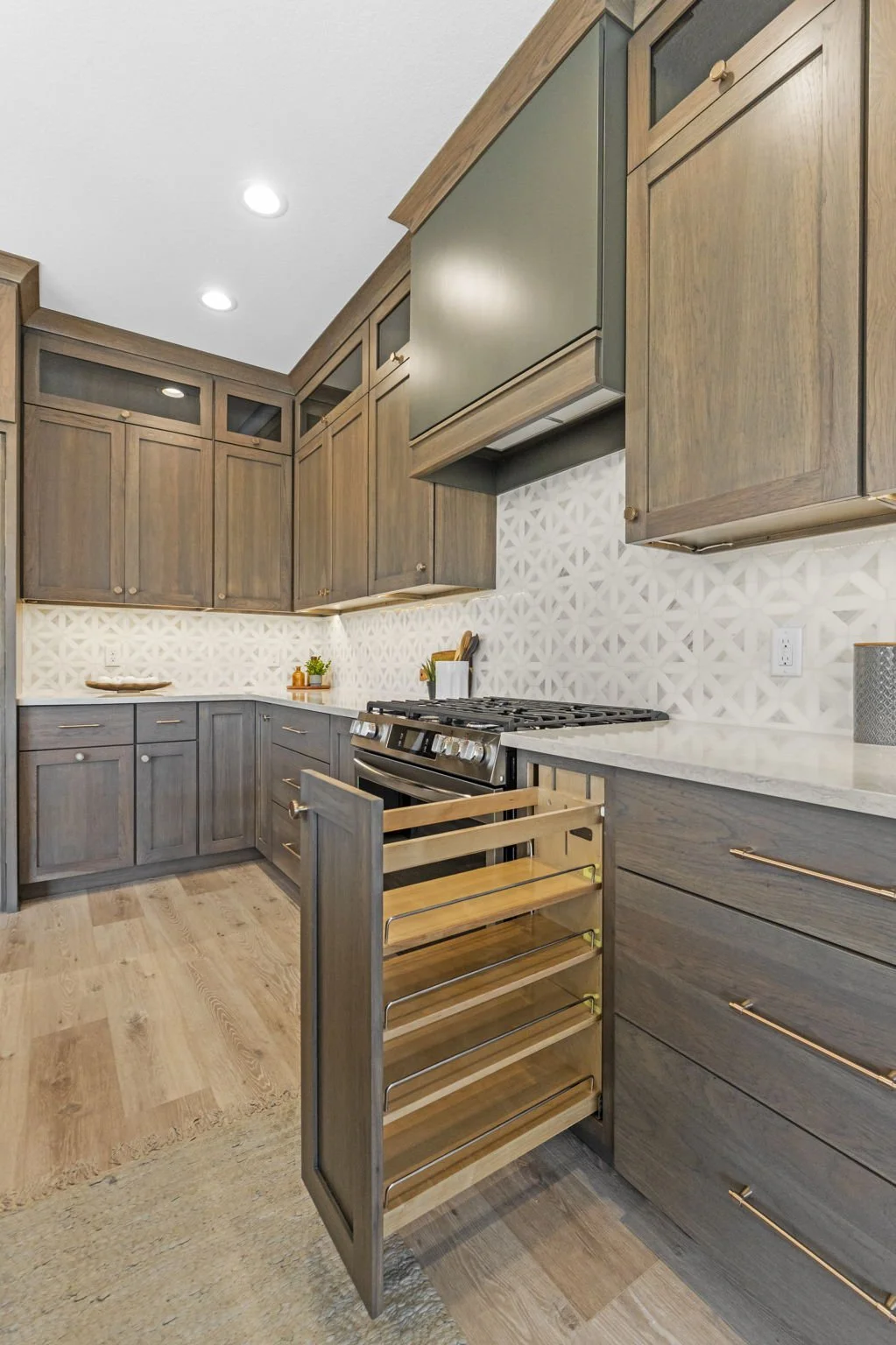 Modern kitchen with wooden cabinets, white patterned backsplash, and a stainless steel stove. A pull-out spice rack is open beside the stove.