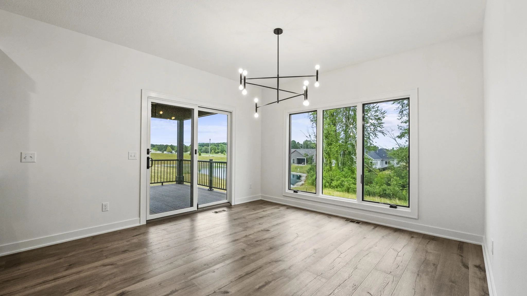 Empty living room with large windows and sliding glass door, hardwood floors, white walls, and modern black chandelier, overlooking a backyard with trees and neighboring houses.