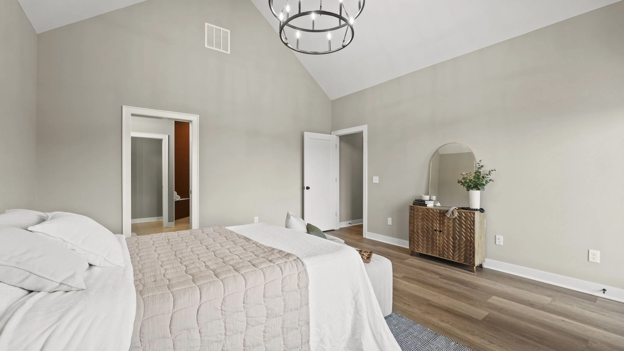 A bedroom with a large bed covered in white and beige bedding, light gray walls, hardwood floors, a round black chandelier hanging from the ceiling, a white door, and a wooden dresser with a mirror and plant on top.