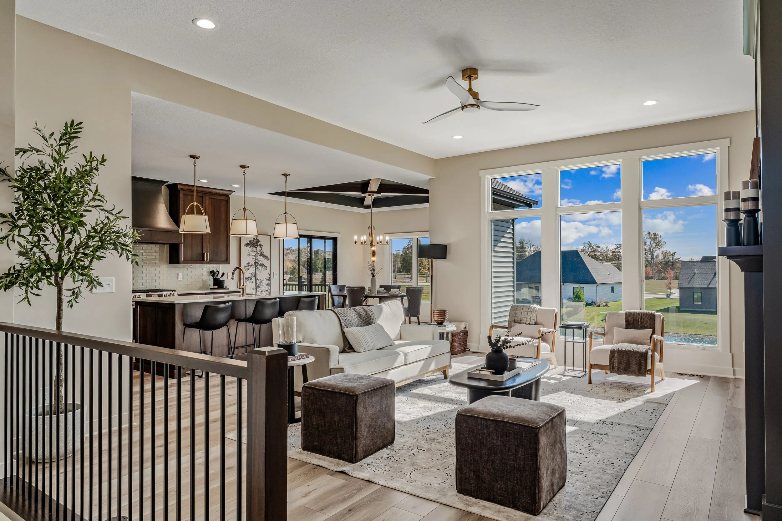 Living room with large windows, white sofa, armchairs, coffee table, ottomans, wood flooring, and kitchen in the background.