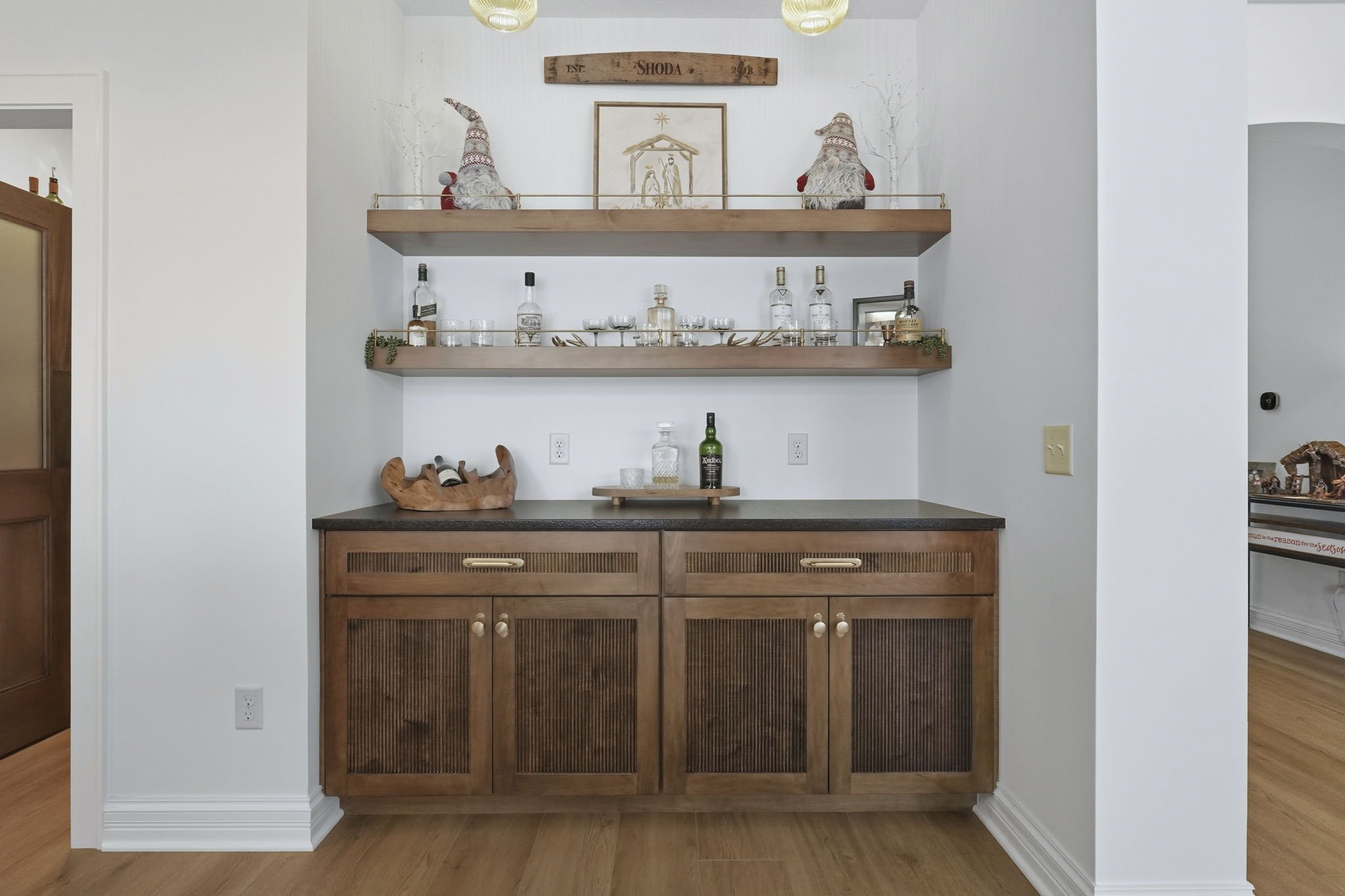 A wooden bar cabinet with two open shelves above it displaying bottles of alcohol, glasses, and decorative items like small Christmas gnomes on top. The wall is white, and the flooring is light wood.