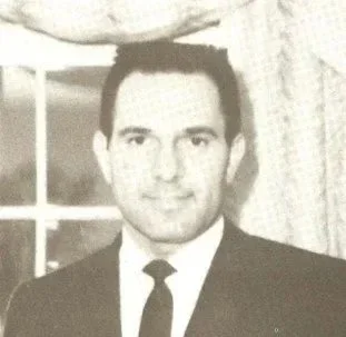 Black and white photo of a man in a suit and tie standing indoors with a window and curtains in the background.