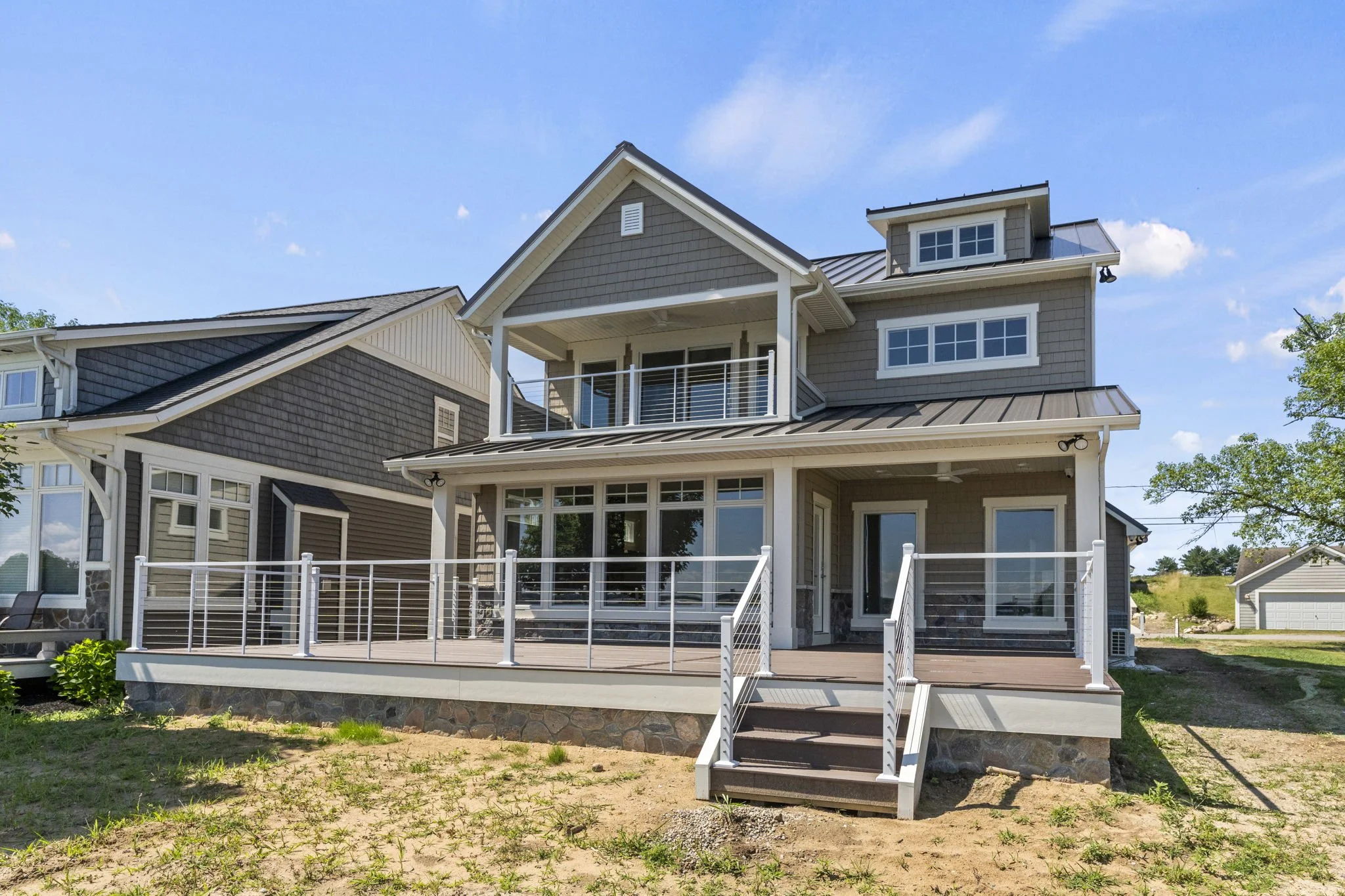 A modern three-story house with a large wooden deck, white railing, multiple windows, and a metal roof, situated under a blue sky.