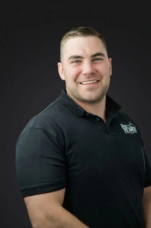 A man smiling, wearing a black polo shirt with a logo on the chest, against a dark background.