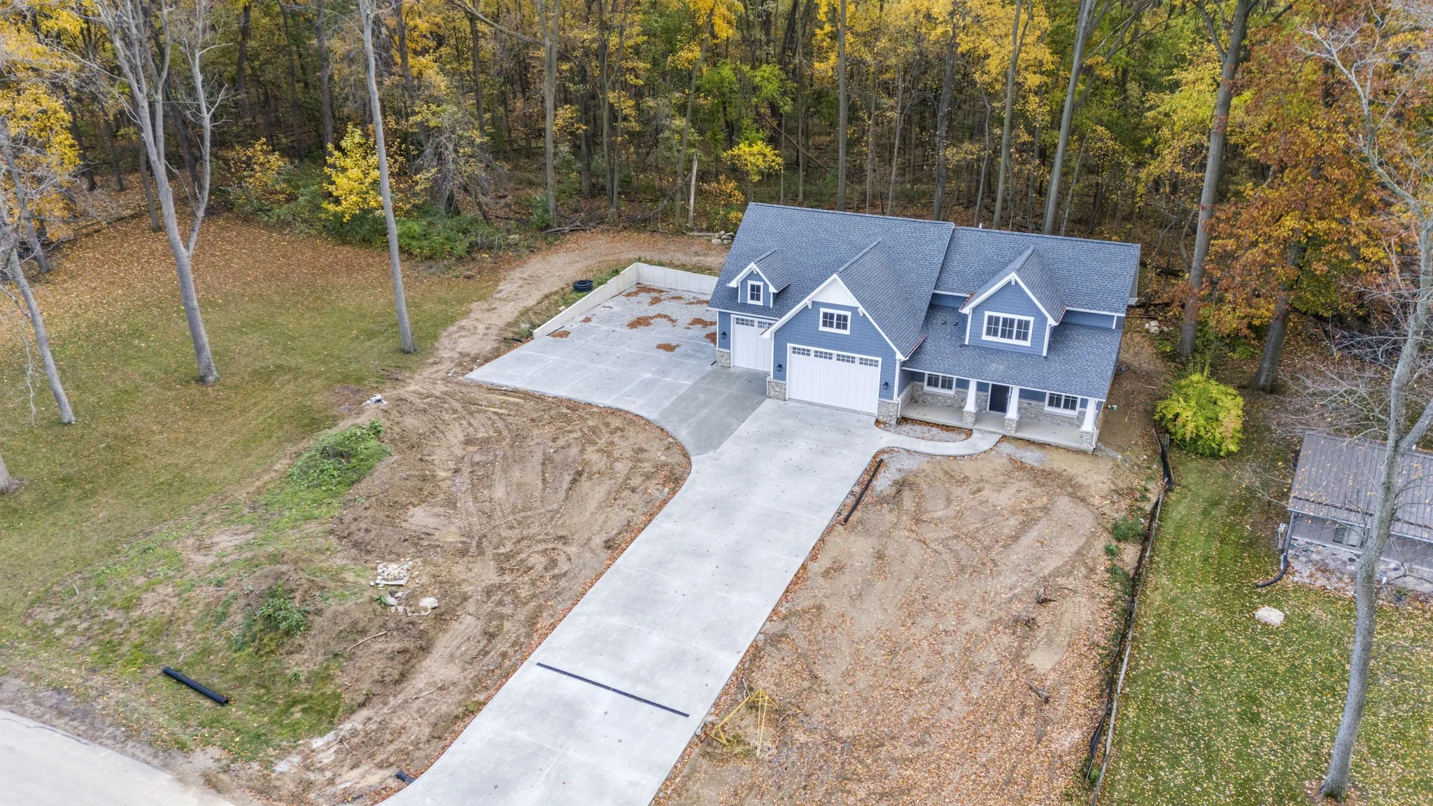 A newly built blue house with a gray roof, surrounded by trees with fall foliage, and a large concrete driveway leading up to the garage. There is some construction activity around the house with dirt and landscaping in progress.