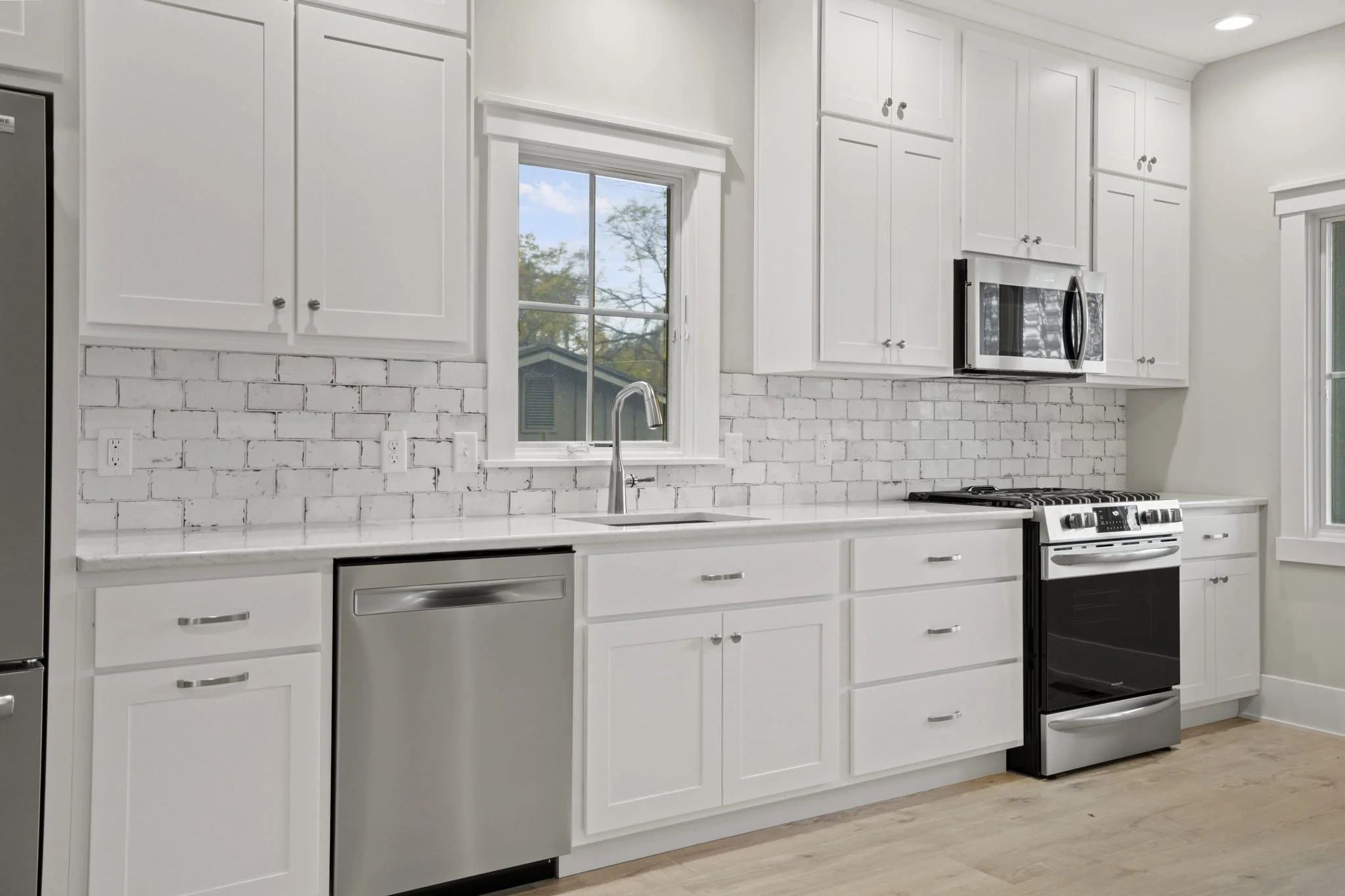 Modern white kitchen with white cabinets, a marble countertop, a white brick backsplash, a double window with trees outside, and stainless steel appliances including a dishwasher, oven, microwave, and refrigerator.