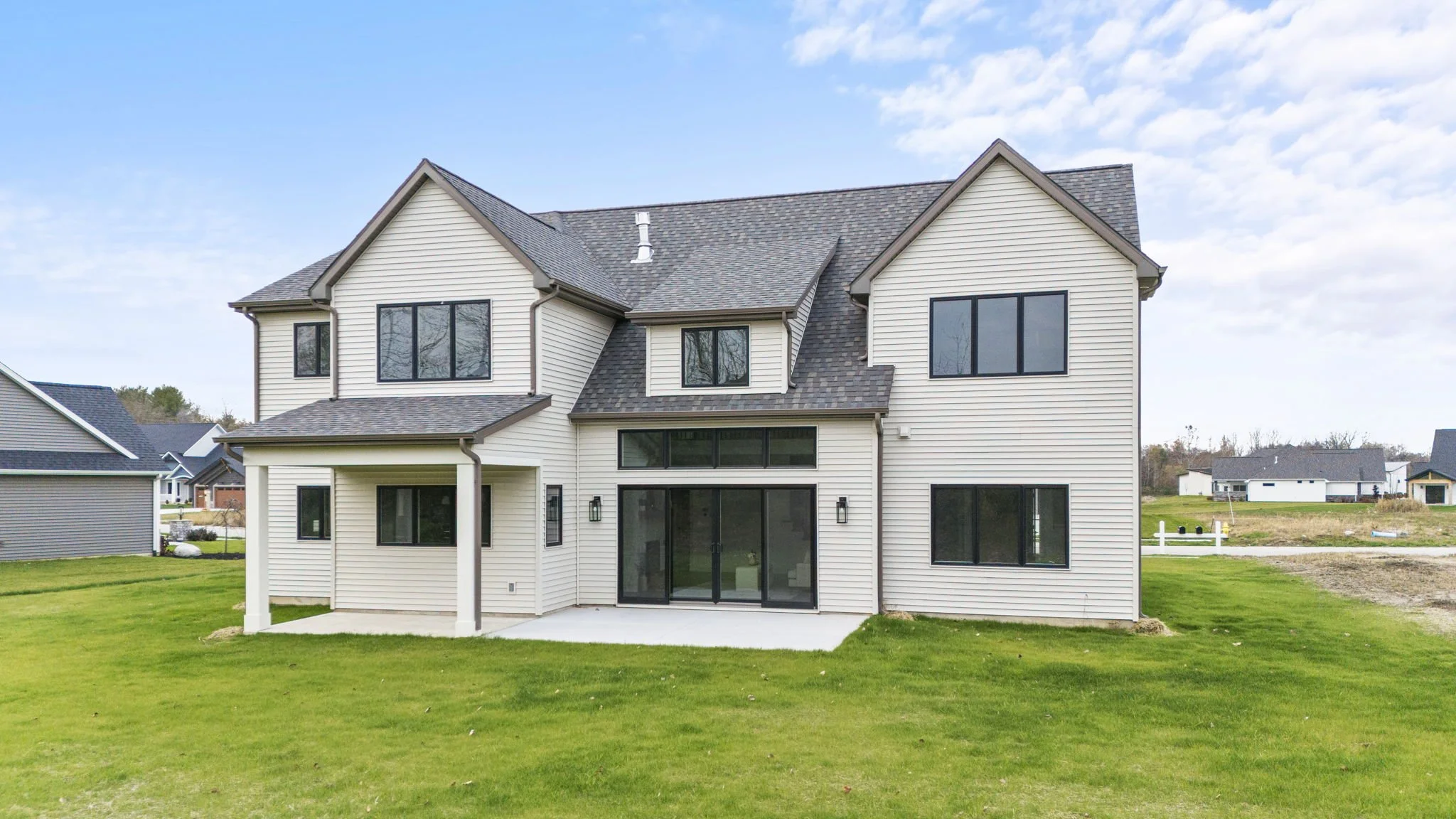 A white two-story house with black windows and a gray shingled roof, located in a grassy yard with a concrete patio at the back and a partly cloudy blue sky overhead.