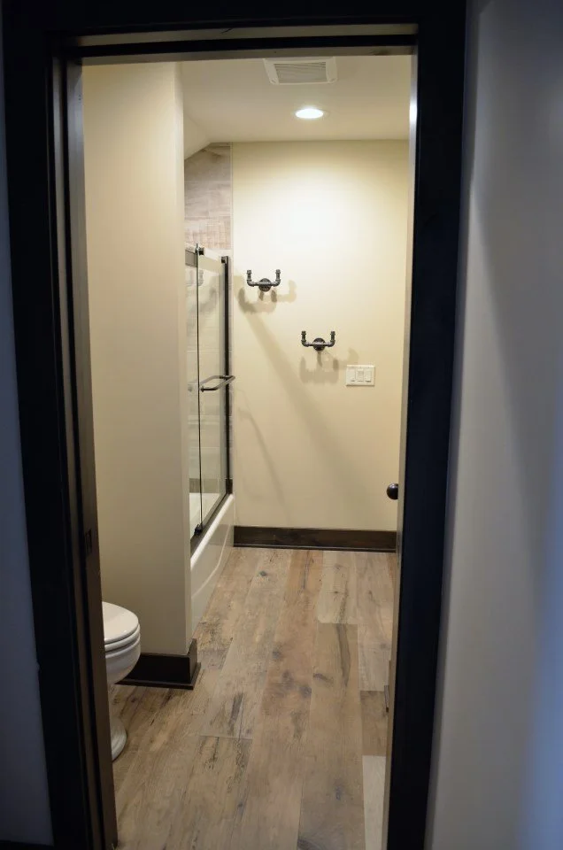 Empty bathroom with a toilet on the left, a glass shower enclosure, and hooks on the back wall, with wood floor and ceiling light.