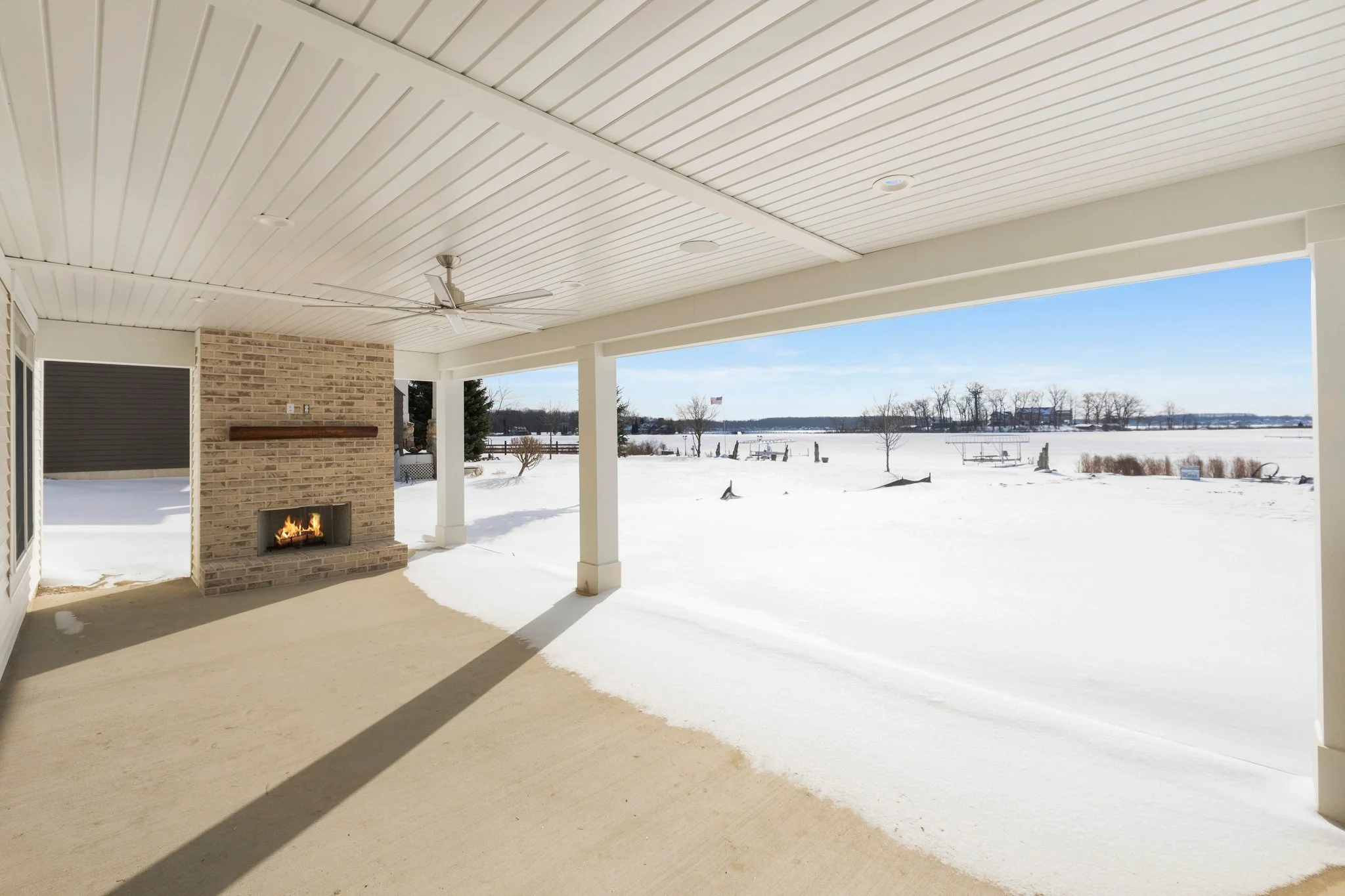 Covered patio with a brick fireplace and a ceiling fan overlooking a snowy landscape with trees and distant buildings.