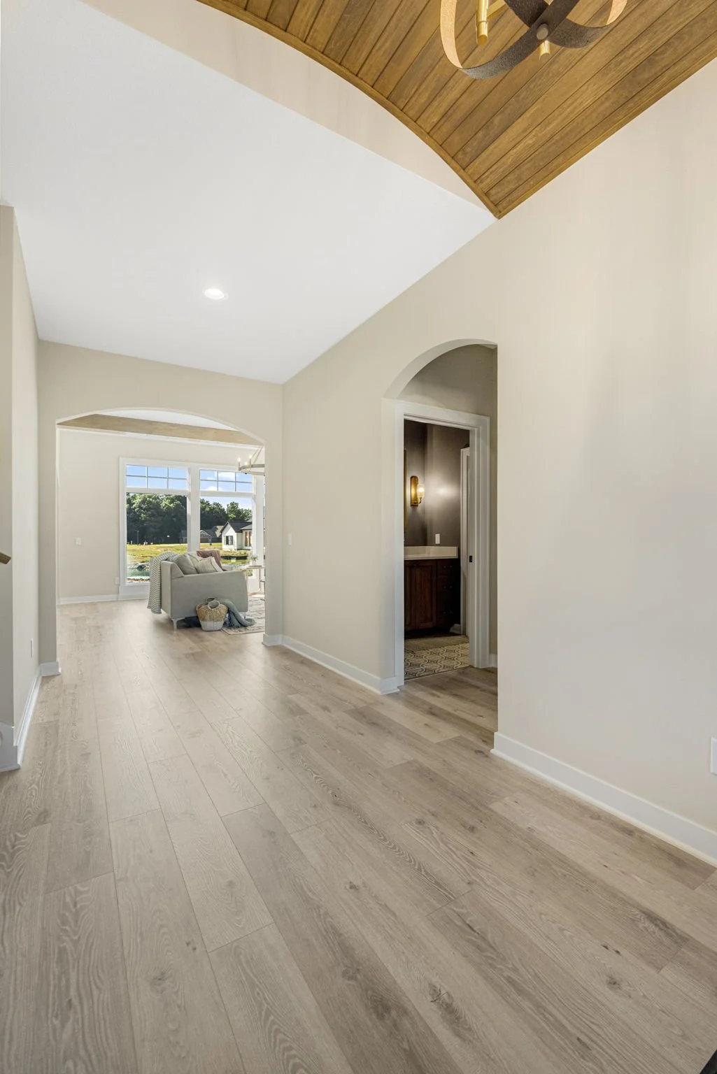 Bright living room with light wooden floors, white walls, large windows, and a cozy seating area near the window, with a view of greenery outside.