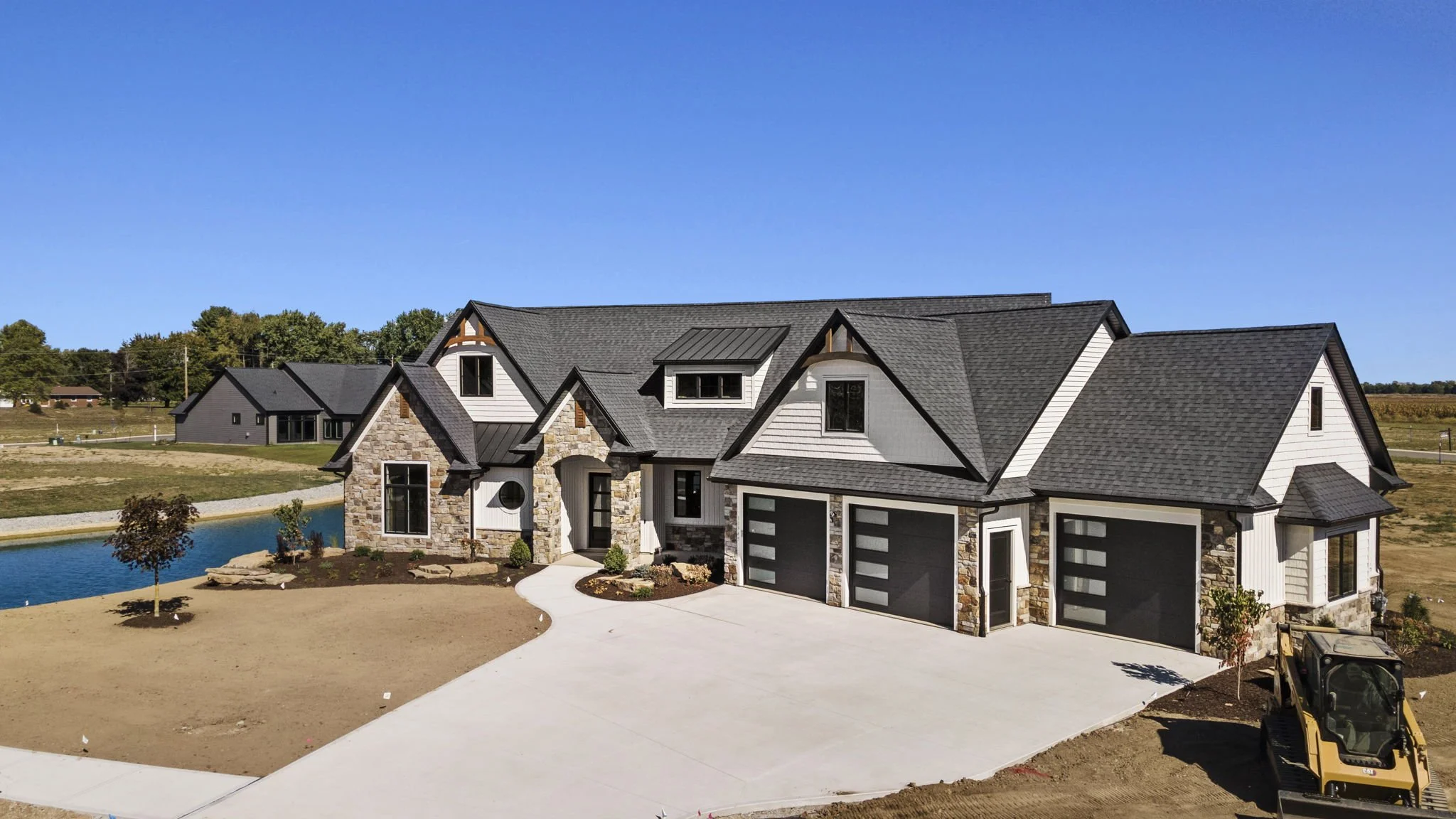 Newly constructed modern house with a stone and white siding exterior, multiple garage doors, and a landscaped yard with a pond, trees, and a small excavator in the driveway under a clear blue sky.