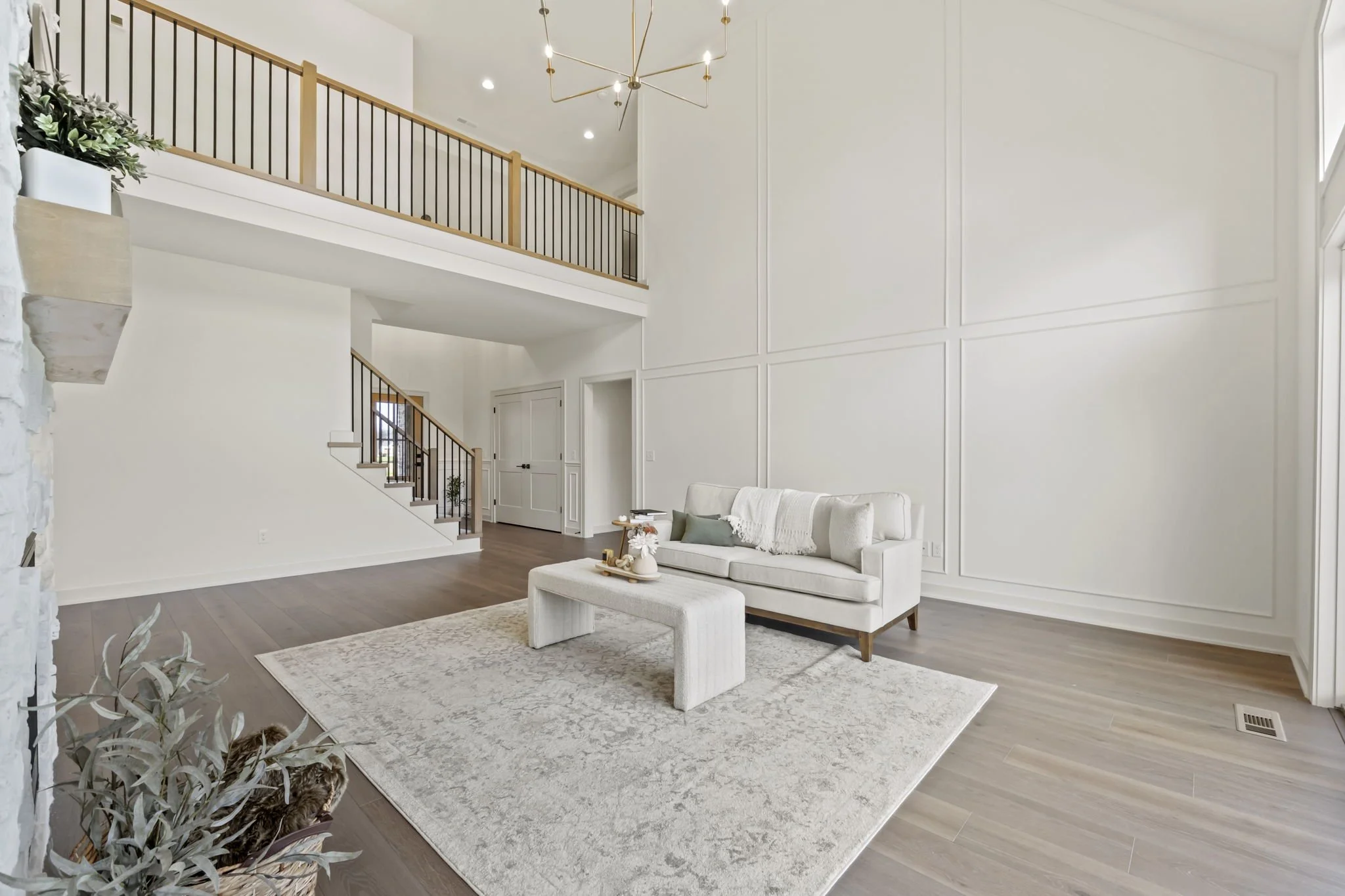 Modern living room with white walls, a white sofa with pillows, a beige area rug, a white upholstered coffee table, and a wooden staircase with black metal railings leading to an upstairs balcony with a wooden railing. Large windows let in natural li